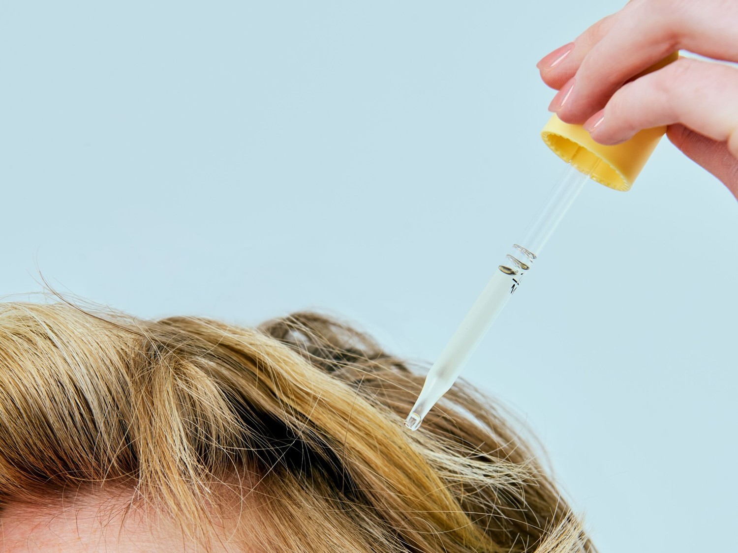 Een hand met lichtroze nagellak houdt een glazen pipet met een gele dop vast, die een transparante vloeistof op blond haar aanbrengt.