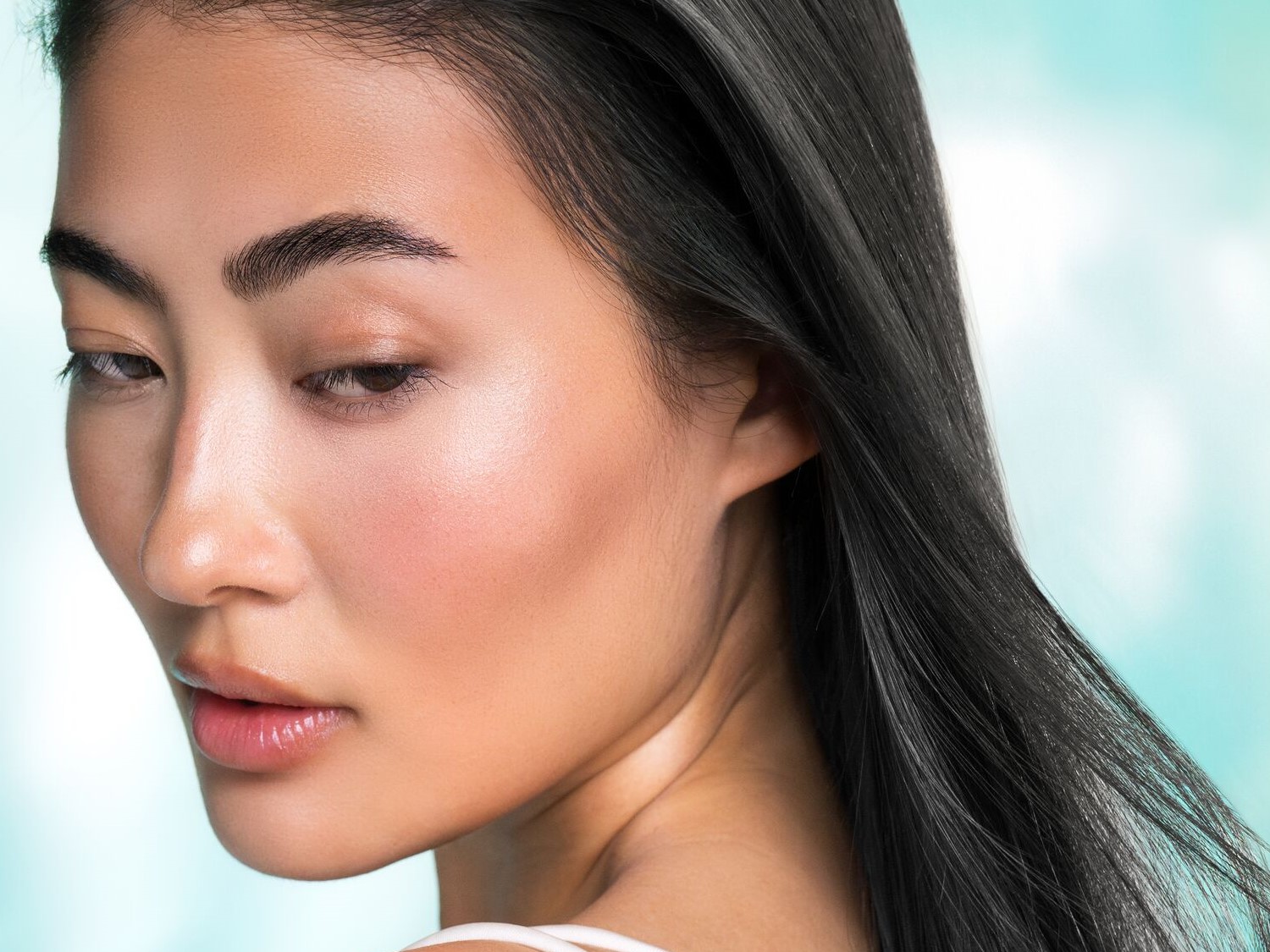 Een close-up van een vrouw met een stralende huid, zachtroze blos op haar wangen en glanzende lippen, haar donkere haar valt over haar schouder.