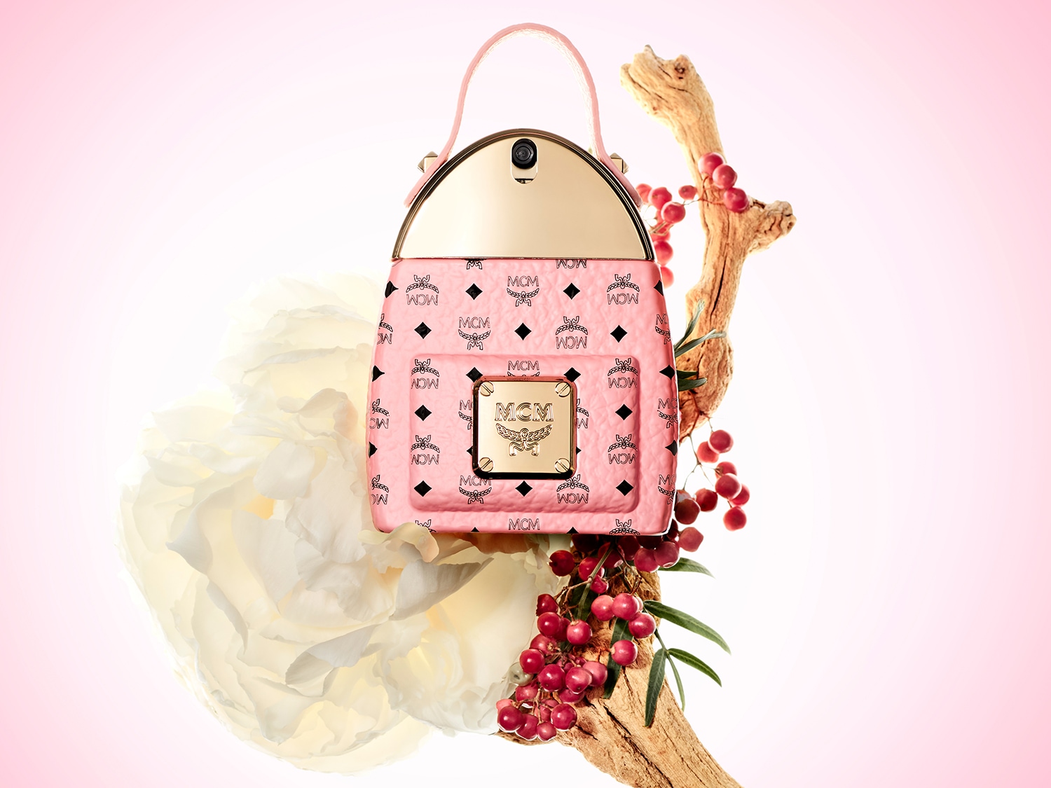 Der MCM Eau de Parfum Flakon in Form eines pinkfarbenen Mini-Rucksacks mit goldenen Akzenten und dem MCM-Logo, umgeben von einer weißen Blüte und roten Beeren, fängt die Essenz von Abenteuer und Romantik ein.