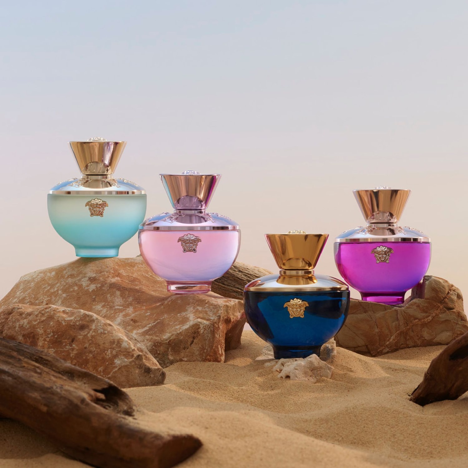 Quatre flacons de parfum Versace, aux couleurs dégradées de bleu clair, rose, bleu foncé et violet, sont posés sur des rochers et du sable, avec des bouchons dorés et le logo Méduse.