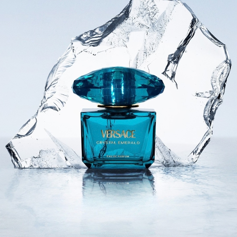 Le parfum Versace Crystal Emerald est présenté dans un flacon rectangulaire en verre teinté de turquoise, avec un bouchon facetté de la même couleur, posé sur une surface réfléchissante avec des éclats de glace en arrière-plan.