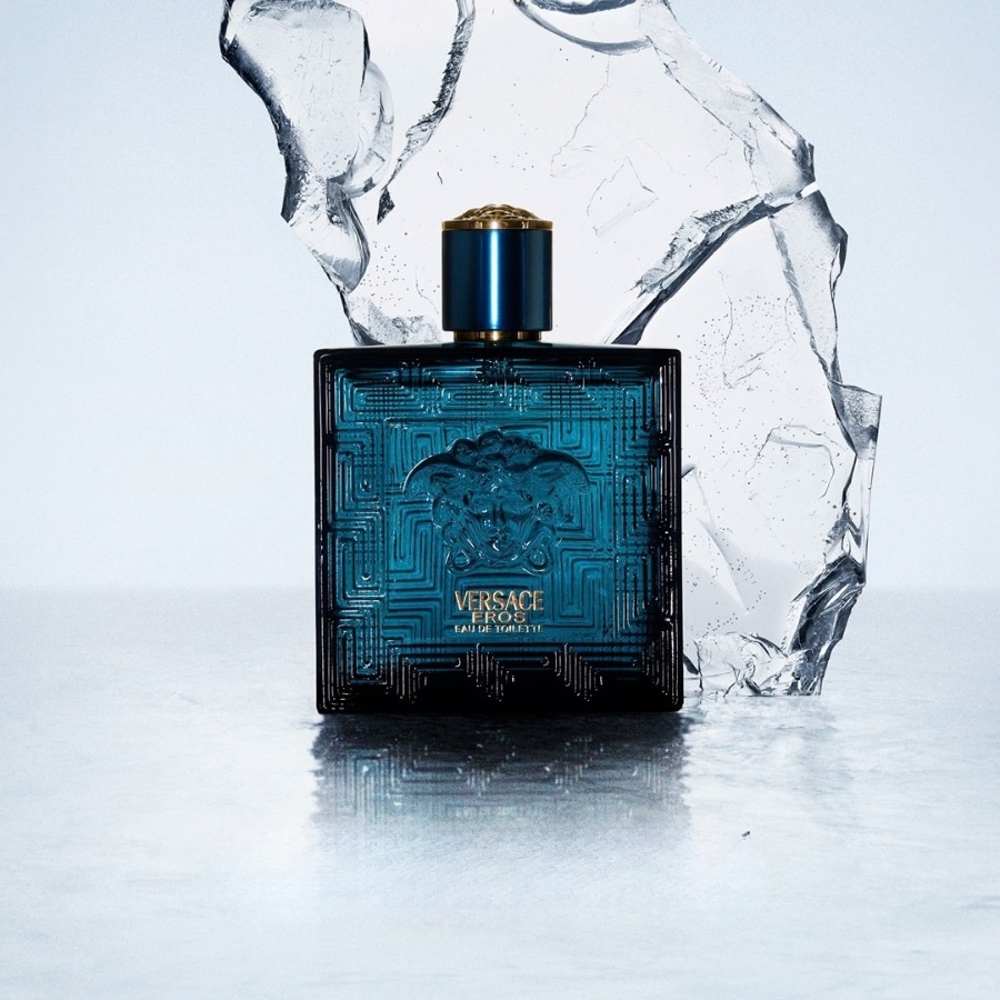 Le parfum Versace Eros Eau de Toilette est présenté dans un flacon rectangulaire bleu sarcelle avec un bouchon doré, posé sur une surface réfléchissante avec des éclats de glace en arrière-plan.