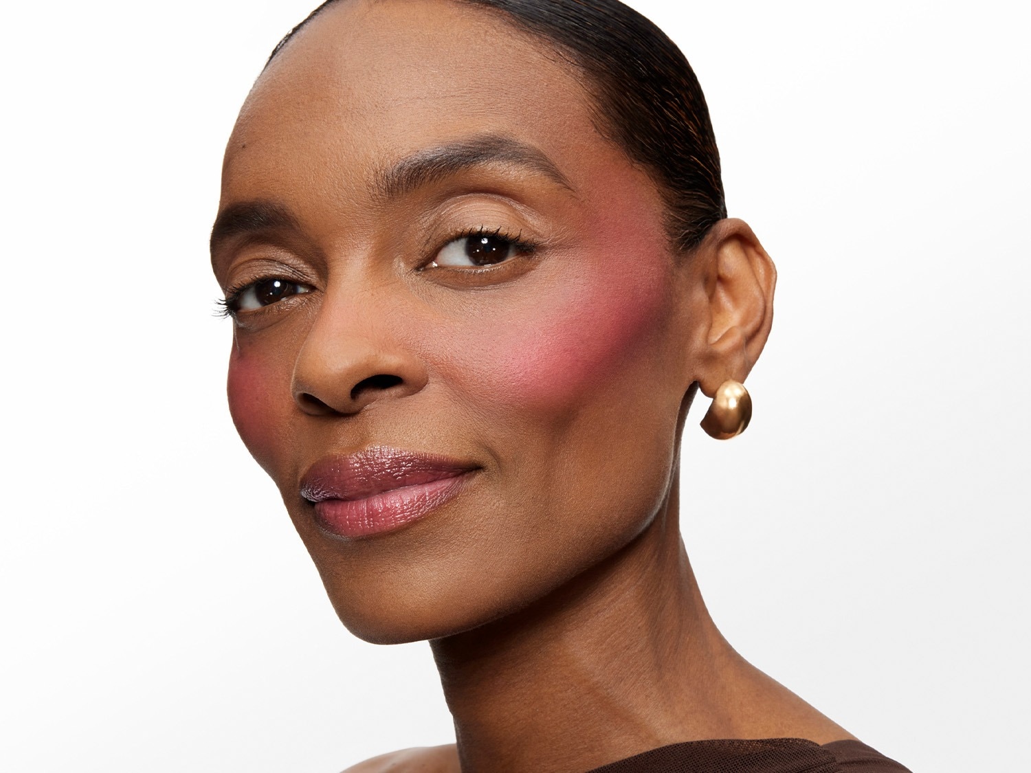 Une femme au teint foncé, les cheveux tirés en arrière, porte un fard à joues rose vif sur les pommettes, un rouge à lèvres brillant et des boucles d'oreilles dorées.