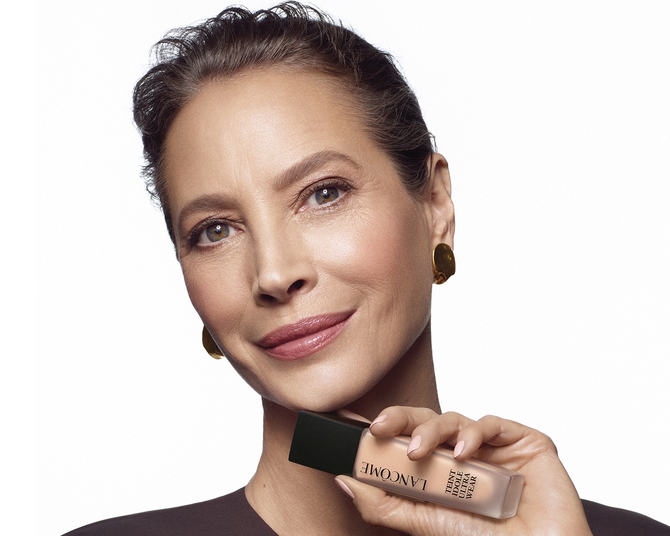 Een vrouw met een stralende huid en een natuurlijke make-up look, houdt een flesje Lancôme Teint Idole Ultra Wear foundation vast, met een zwarte dop en een matte glazen flesje dat de lichte beige kleur van de foundation onthult.