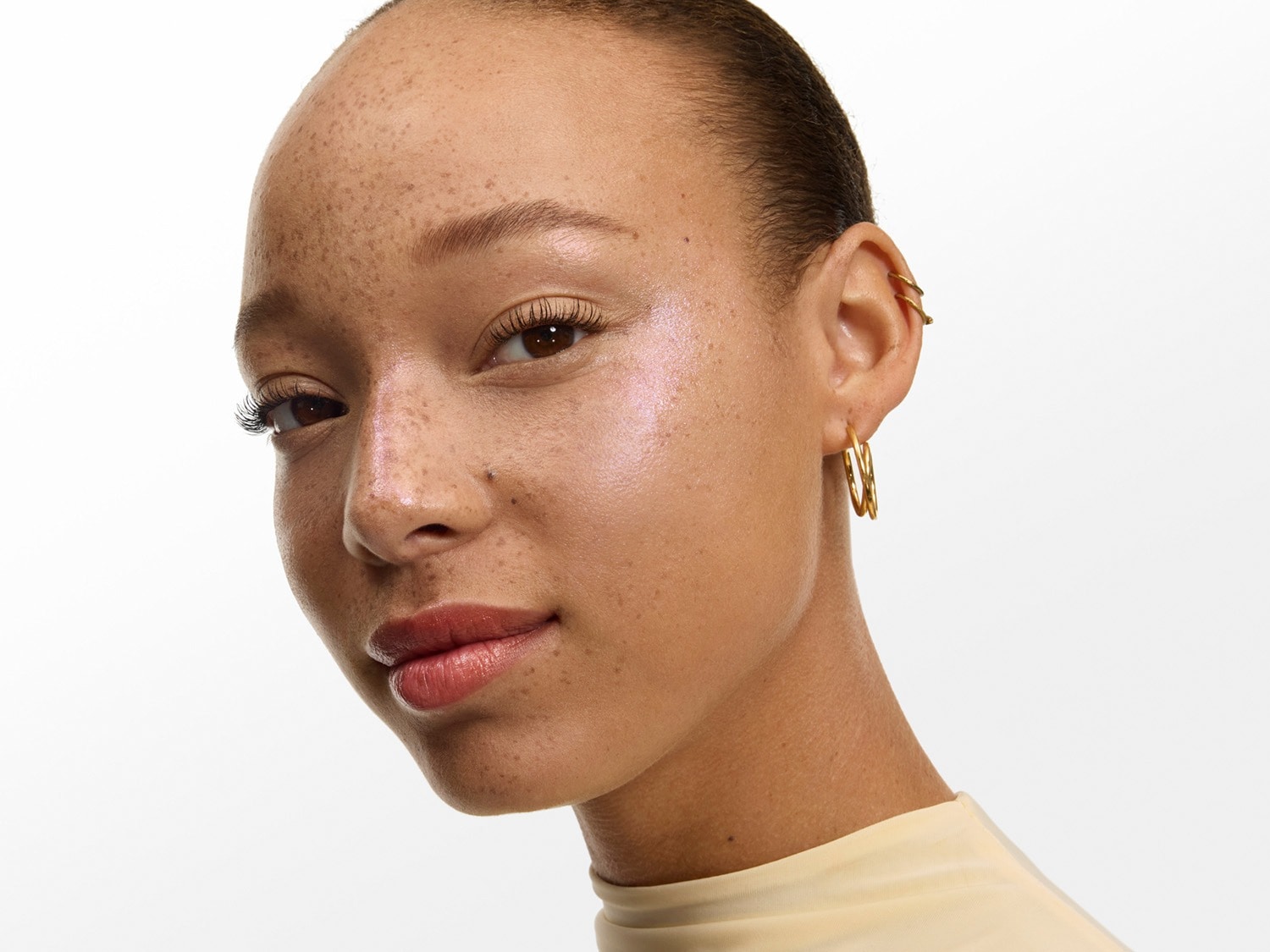 Une femme au teint clair et aux taches de rousseur porte un maquillage lumineux, avec un fard à joues irisé et un rouge à lèvres brillant, complété par des boucles d'oreilles créoles dorées et un piercing d'oreille.