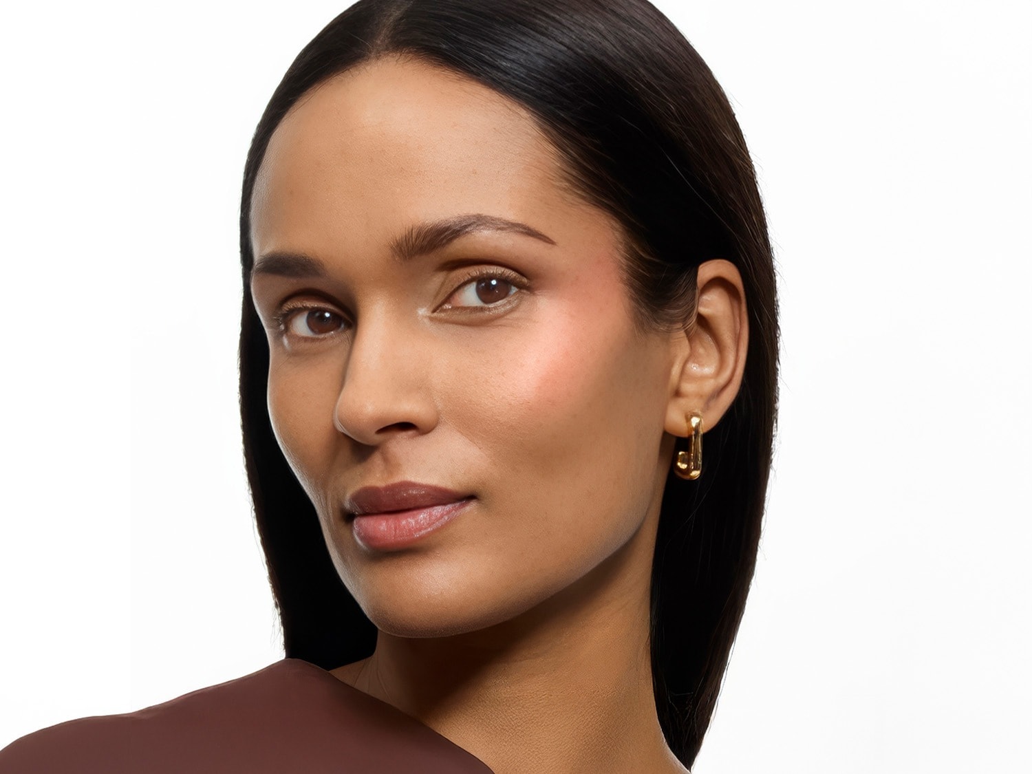 Une femme à la peau mate et aux cheveux noirs porte un fard à joues rose clair sur les pommettes, un rouge à lèvres brillant et des boucles d'oreilles dorées en forme de J.