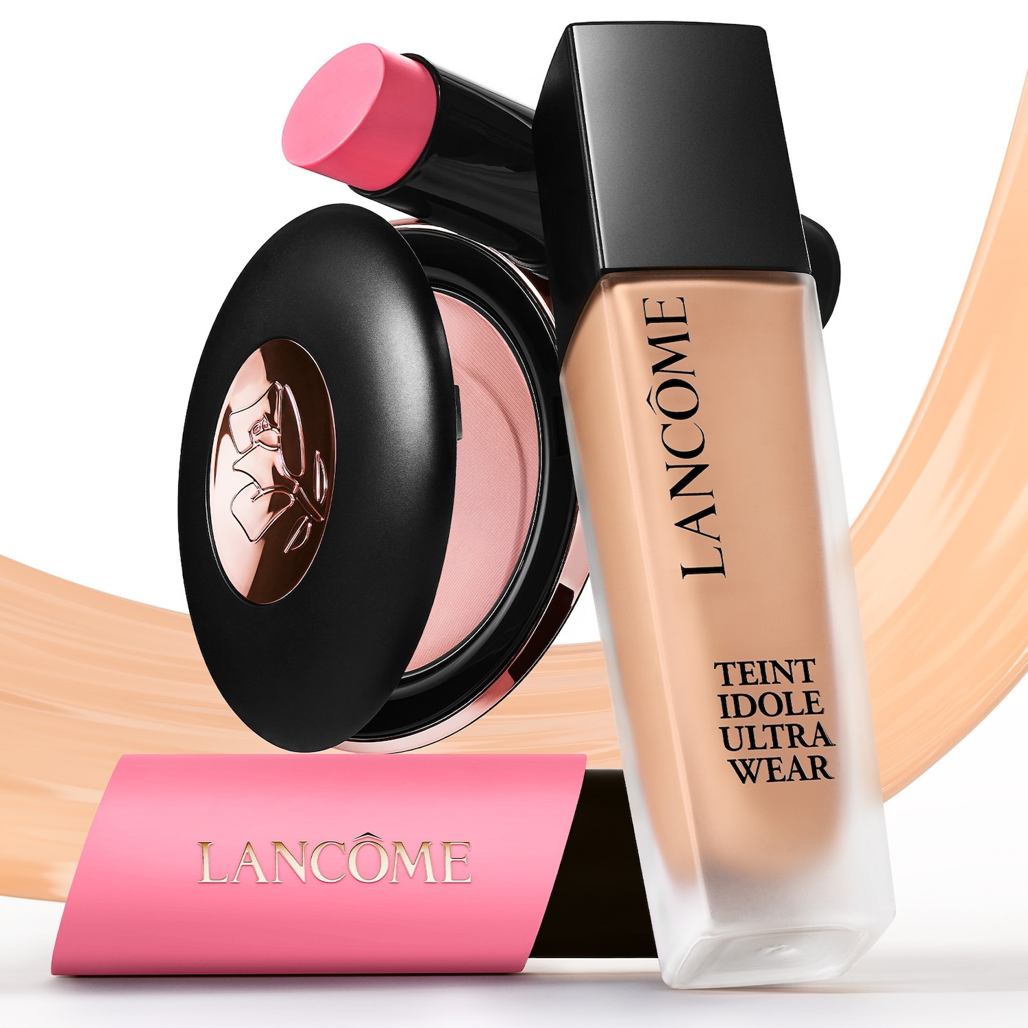 Een Lancôme Teint Idole Ultra Wear foundationflesje met een zwarte dop, een roze blushstick en een compacte poederdoos met een roze blush, alles tegen een witte achtergrond met een roze Lancôme-logo.