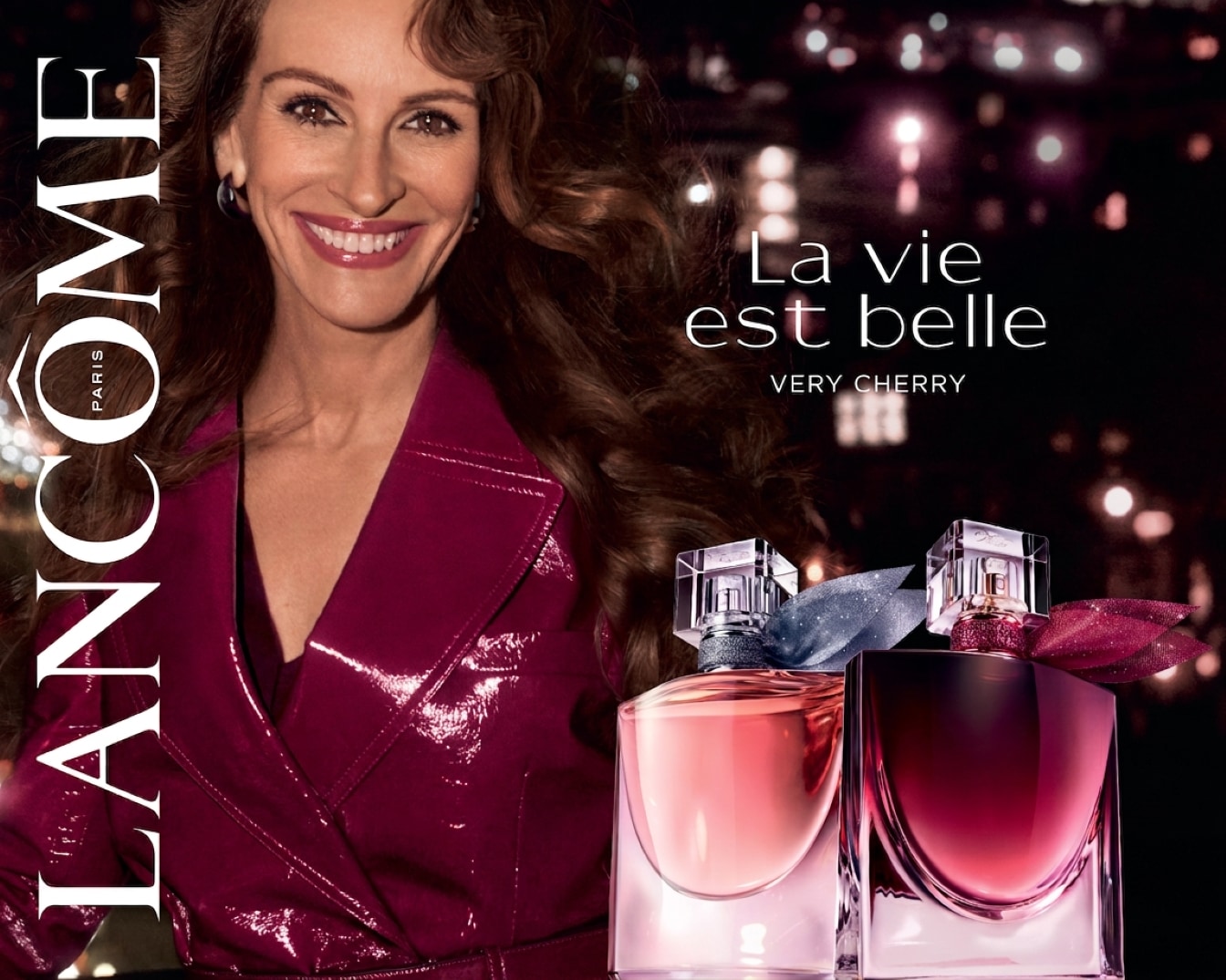 Julia Roberts lächelt in einer glänzenden, weinroten Jacke, während sie zwei Flaschen Lancôme La vie est belle Very Cherry Parfüm präsentiert, eine in hellem Rosa und die andere in dunklem Weinrot, beide mit glitzernden Bändern verziert.