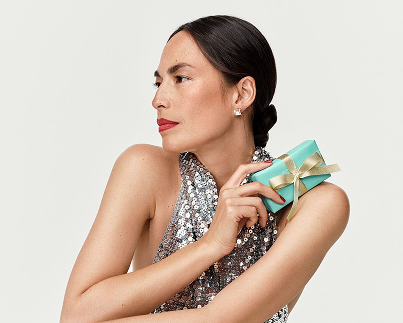 Žena s uhlazenými vlasy, výraznými červenými rty a třpytivými stříbrnými šaty drží tyrkysovou dárkovou krabičku se zlatou stuhou, zatímco se dívá do strany.