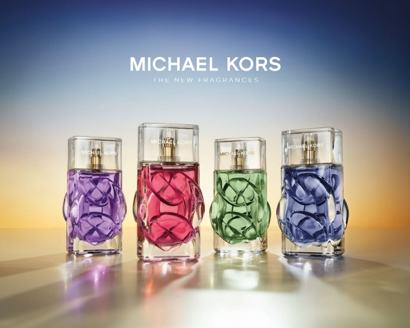 Vier Michael Kors parfums in paars, roze, groen en blauw, elk in een transparante fles met een uniek, golvend ontwerp en een gouden dop, staan op een glanzend oppervlak tegen een achtergrond die van blauw naar oranje verloopt.