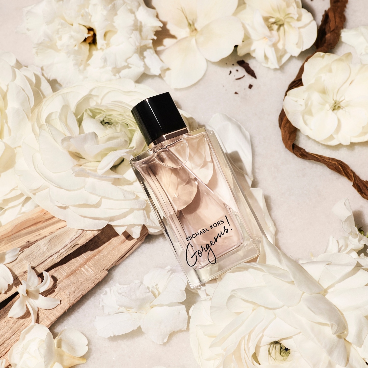 Le parfum Michael Kors Gorgeous est présenté dans un flacon rectangulaire en verre transparent avec un bouchon noir, posé sur un lit de fleurs blanches et de copeaux de bois, évoquant une ambiance florale et boisée.