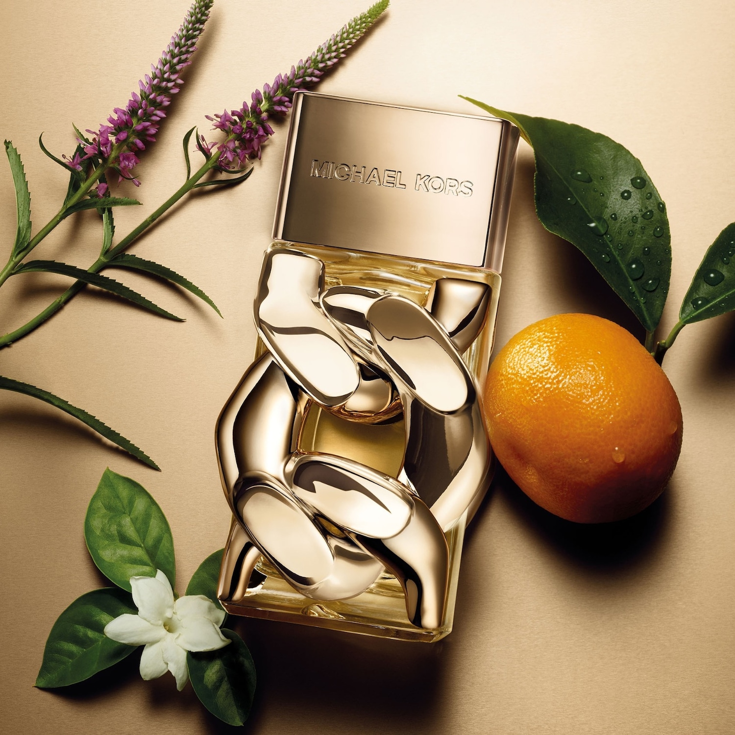 Le parfum Michael Kors est présenté dans un flacon en verre transparent avec des maillons de chaîne dorés, posé sur une surface dorée, entouré de fleurs violettes, de feuilles vertes et d'une orange.