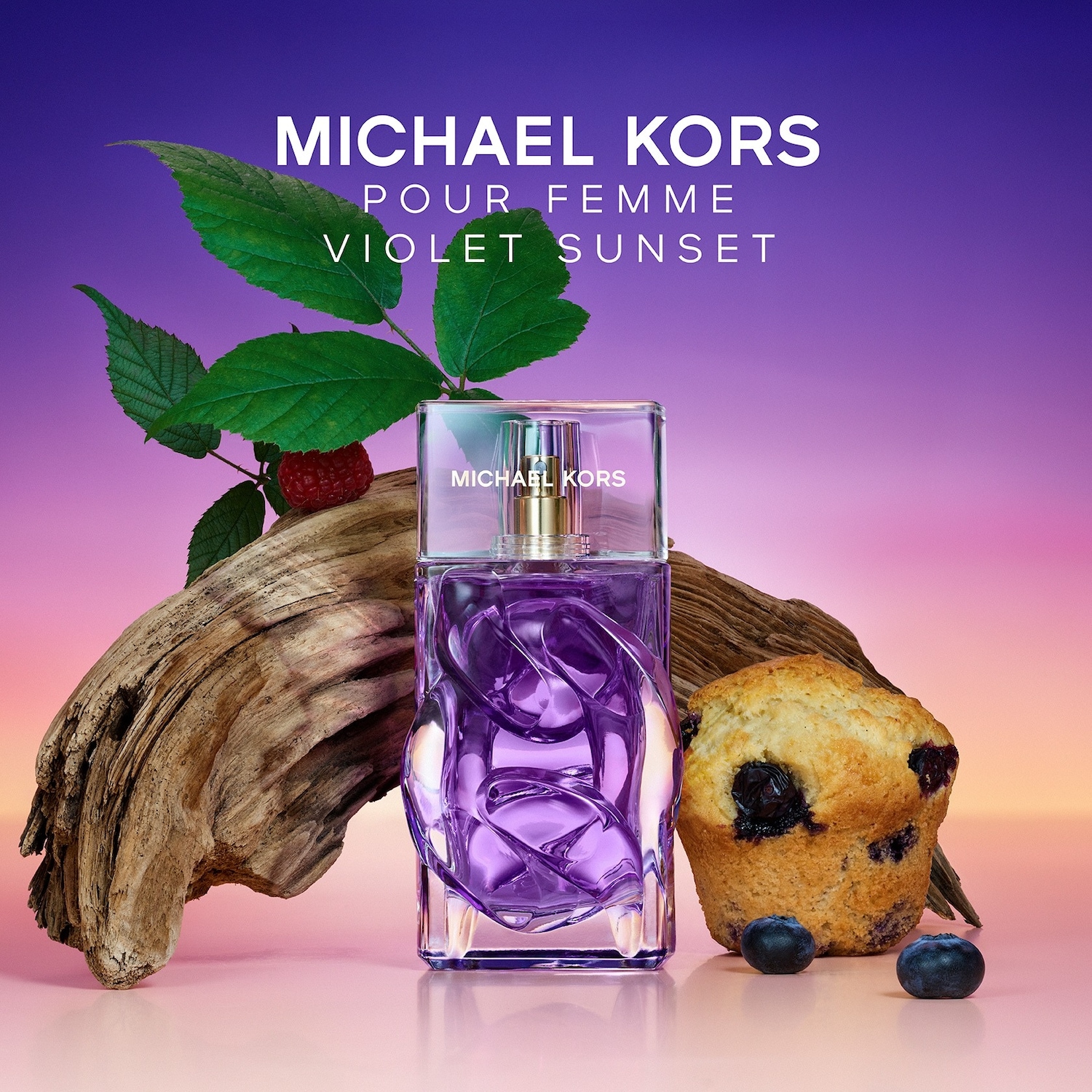 Le parfum Michael Kors Pour Femme Violet Sunset est présenté dans un flacon en verre violet avec des motifs tourbillonnants, posé à côté d'un morceau de bois flotté, d'une framboise, de myrtilles et d'un muffin aux myrtilles, sur un fond dégradé violet et rose.