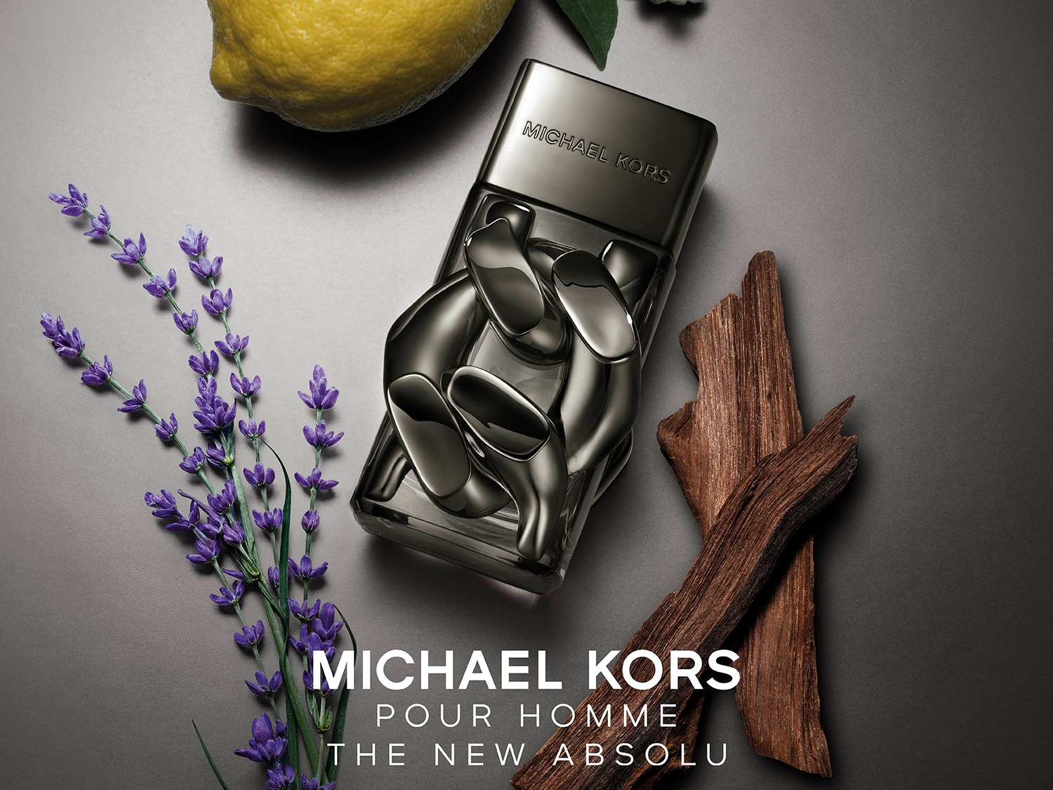 Le parfum Michael Kors Pour Homme The New Absolu est présenté dans un flacon en verre transparent avec un bouchon noir, entouré de lavande violette, d'un citron jaune et de morceaux de bois, sur un fond gris foncé.