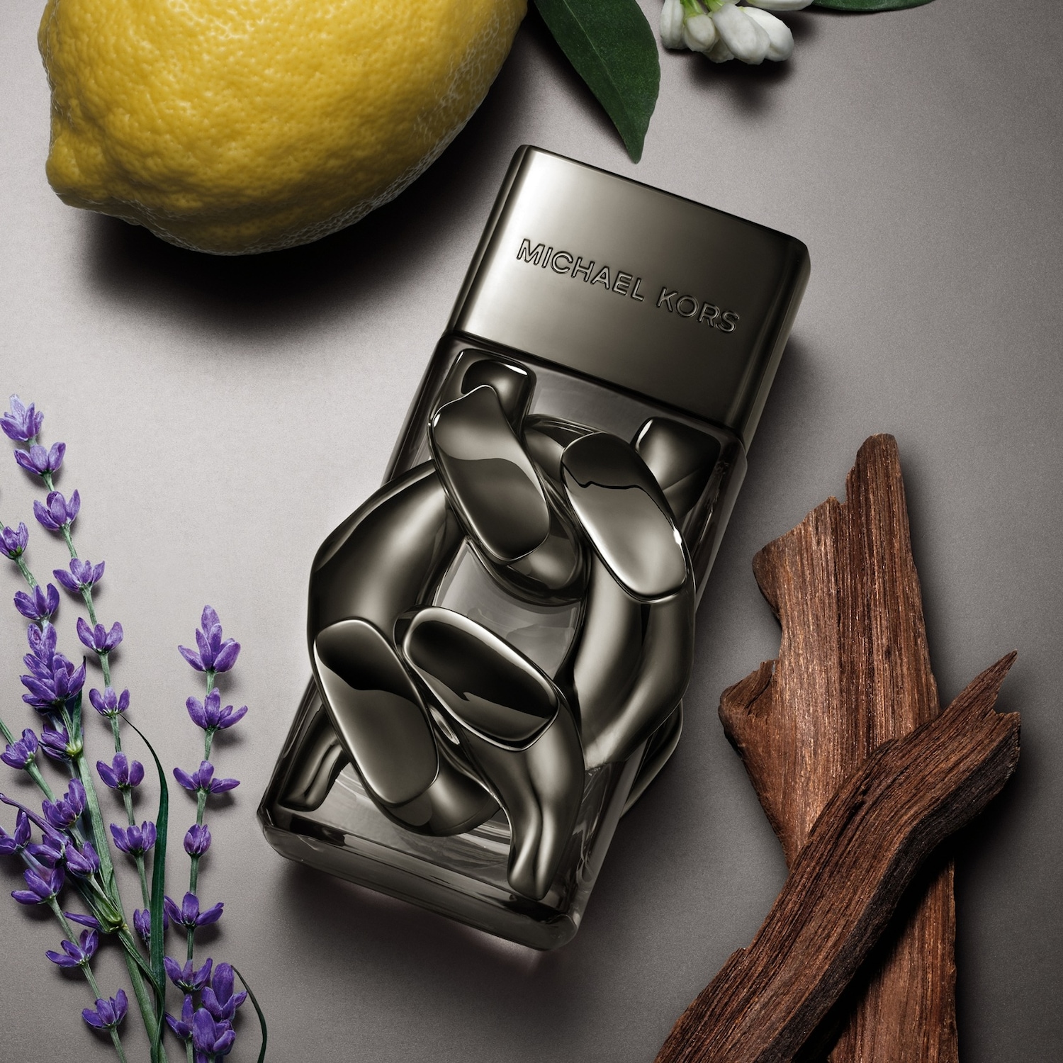 Le parfum Michael Kors pour homme est présenté dans un flacon en verre gris foncé avec un bouchon assorti, entouré d'un citron jaune vif, de lavande violette, de fleurs blanches et de morceaux de bois, le tout sur un fond gris.