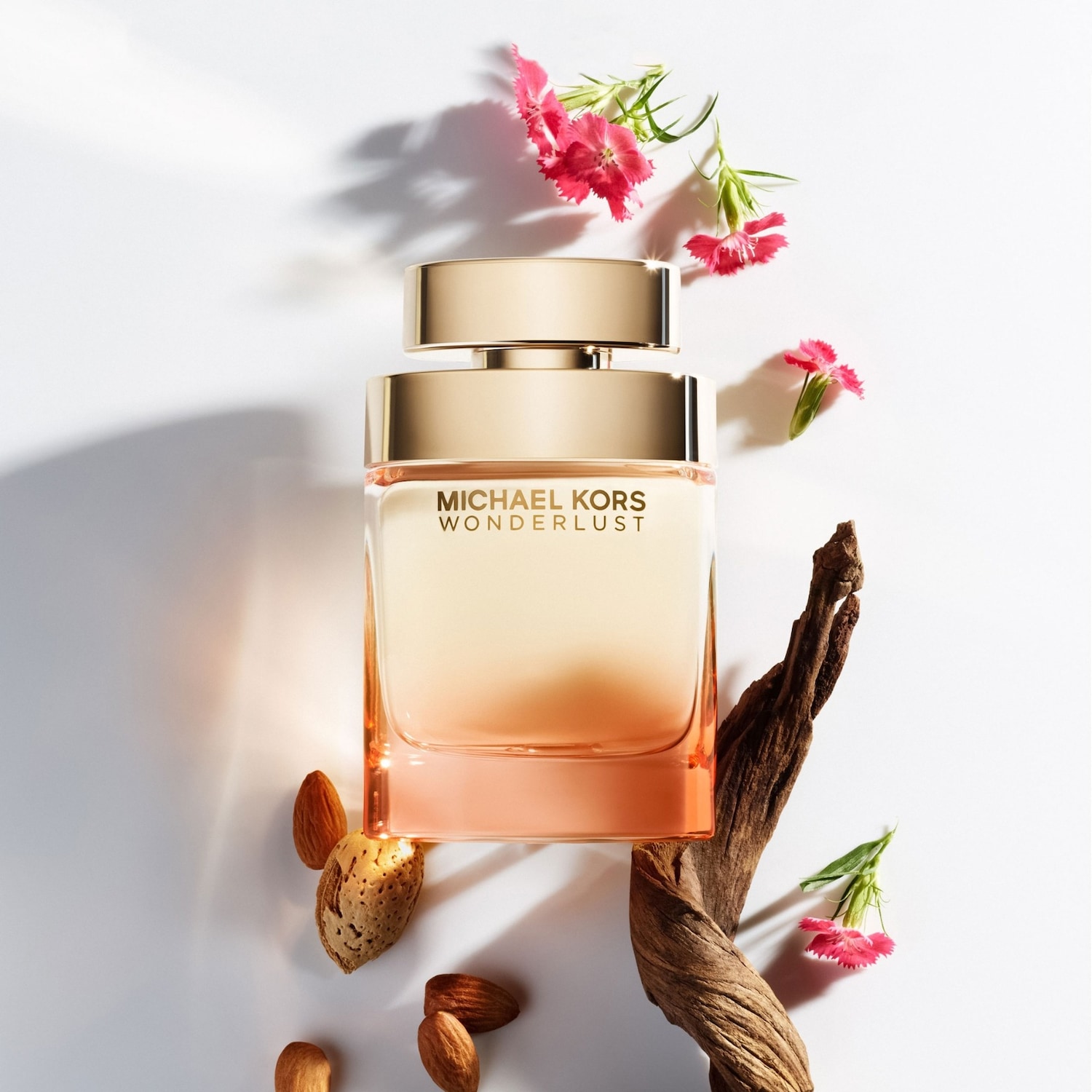Le parfum Michael Kors Wonderlust est présenté dans un flacon rectangulaire en verre, avec un dégradé de couleurs allant du pêche clair au corail, surmonté d'un bouchon doré, entouré d'amandes, de bois flotté et de petites fleurs roses.