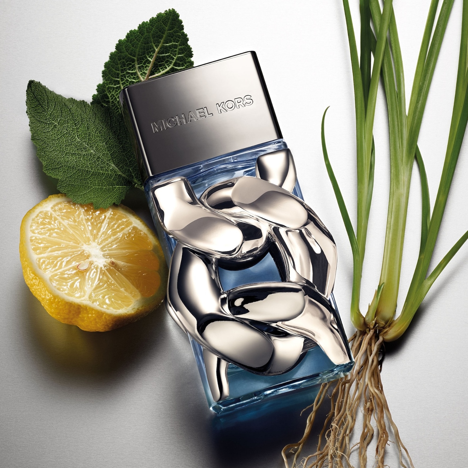 Le parfum Michael Kors pour homme est présenté dans un flacon rectangulaire bleu avec un motif de chaîne argentée, accompagné d'une demi-tranche de citron, de feuilles de sauge et d'une plante verte avec des racines, le tout sur un fond gris clair.