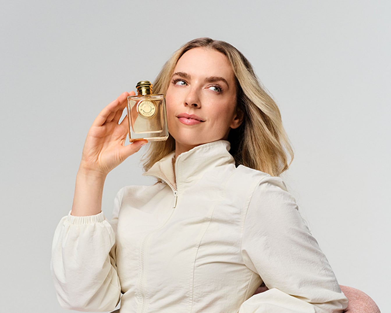 Eine Frau mit blonden Haaren und einem beigefarbenen Oberteil hält einen rechteckigen Parfümflakon mit goldener Kappe und goldenem Etikett in der Hand und blickt nach oben.