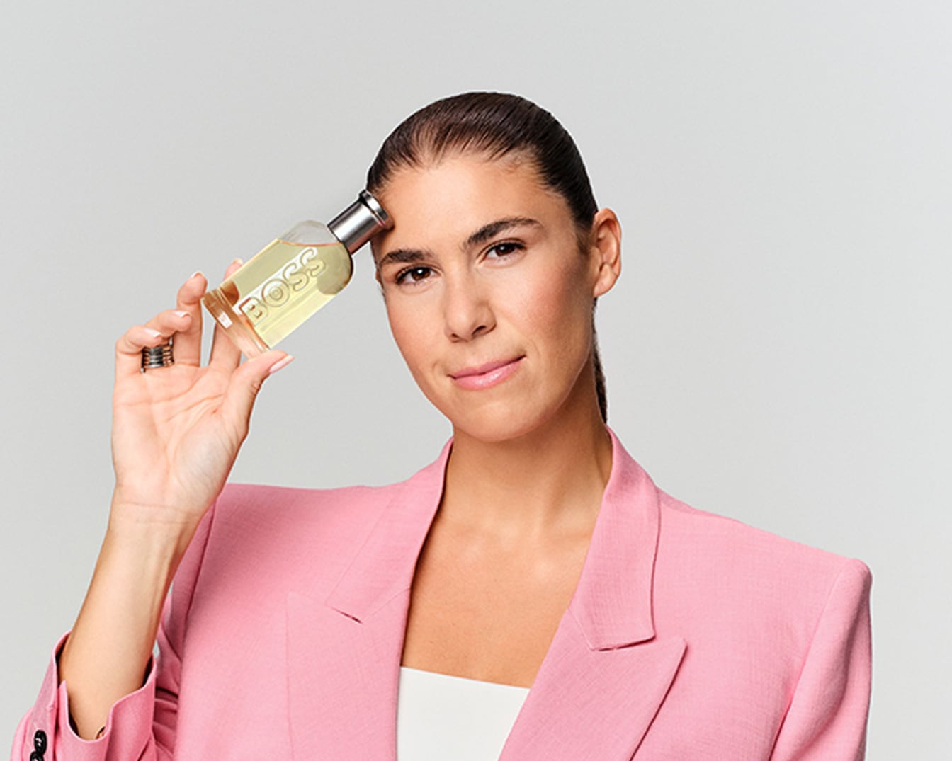 Eine Frau mit einem rosafarbenen Blazer hält einen rechteckigen BOSS-Parfümflakon mit goldener Flüssigkeit und silbernem Verschluss an ihre Schläfe.