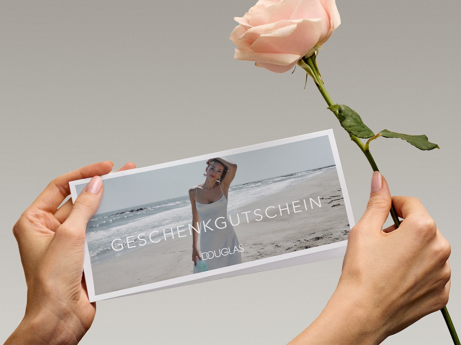 Zwei Hände halten eine Geschenkkarte mit der Aufschrift "GESCHENKGUTSCHEIN" und dem Douglas-Logo, während eine Hand zusätzlich eine einzelne rosa Rose hält.