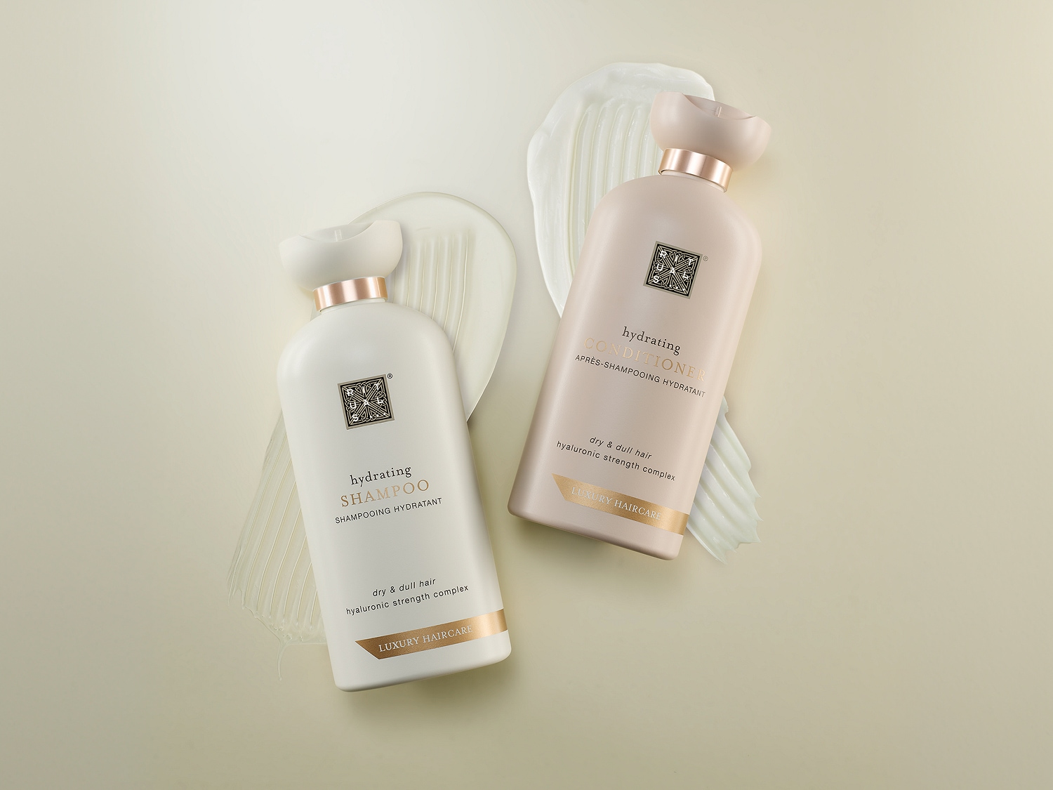 Zwei luxuriöse Haarpflegeprodukte in mattweißen Flaschen mit rosé-goldenen Akzenten, platziert auf cremigem Schaum und Texturen, für trockened und glanzloses Haar.