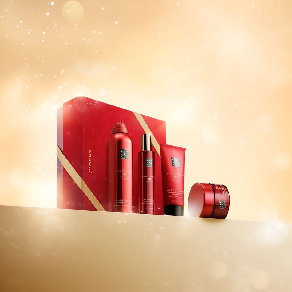 Das Rituals Geschenkset in Rot und Gold enthält vier Produkte: einen Duschschaum, ein Haar- und Körperspray, eine Körpercreme und ein Peeling, die alle auf einem goldenen Podest vor einem verschwommenen, warmen Hintergrund mit funkelnden Lichtern stehen.