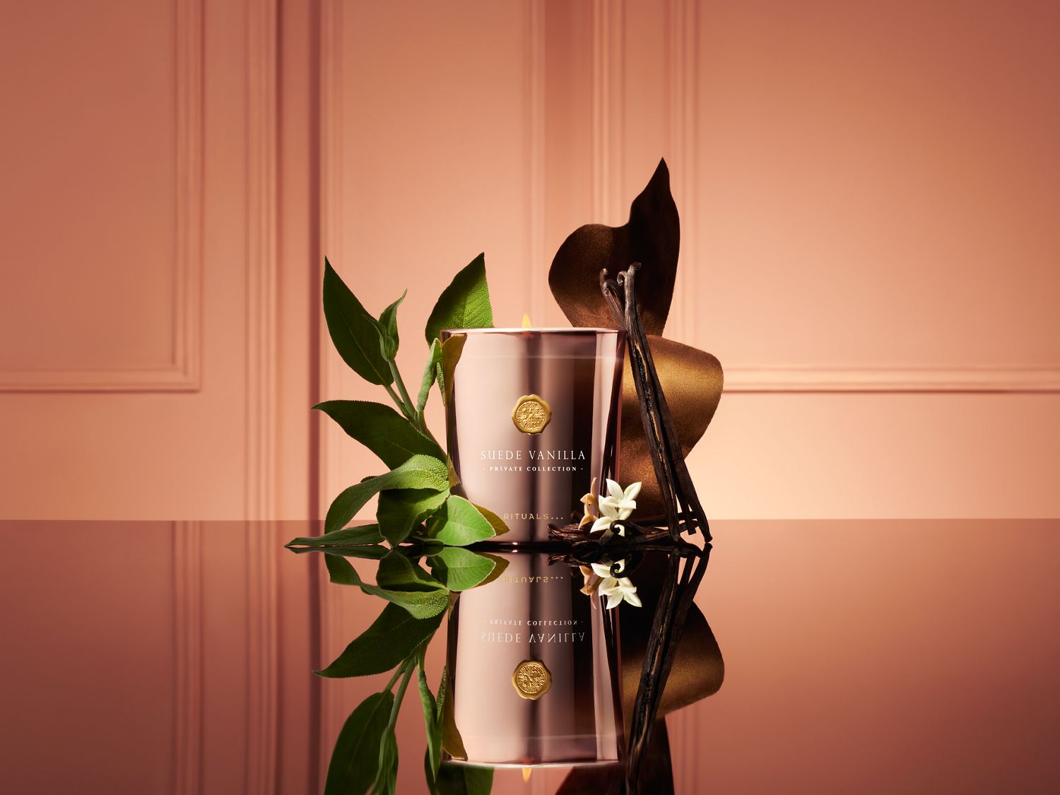 Eine elegante Suede Vanilla Duftkerze in einem kupferfarbenen Glas aus der Private Collection von Rituals, dekorativ arrangiert mit grünen Blättern und Vanilleschoten vor einem warmen Hintergrund.