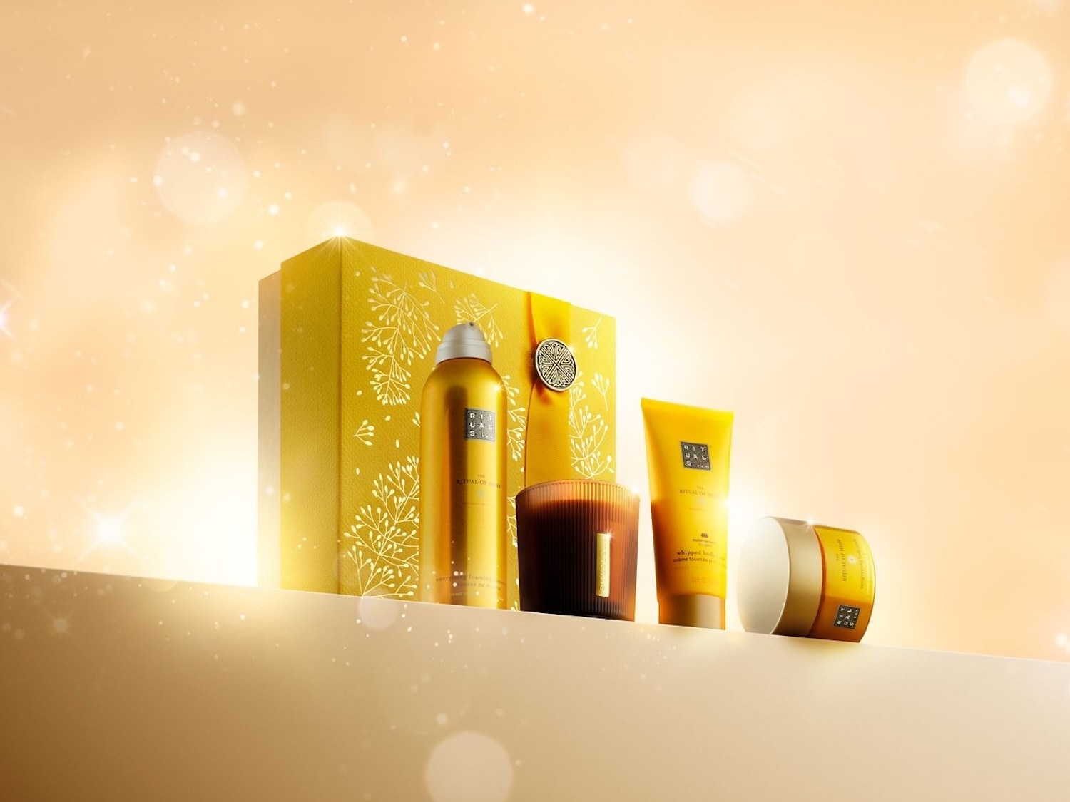 Ein strahlendes Geschenkset von Rituals mit Duschschaum, Duftkerze, Körpercreme und Körperpeeling vor einem festlich goldenen Hintergrund.