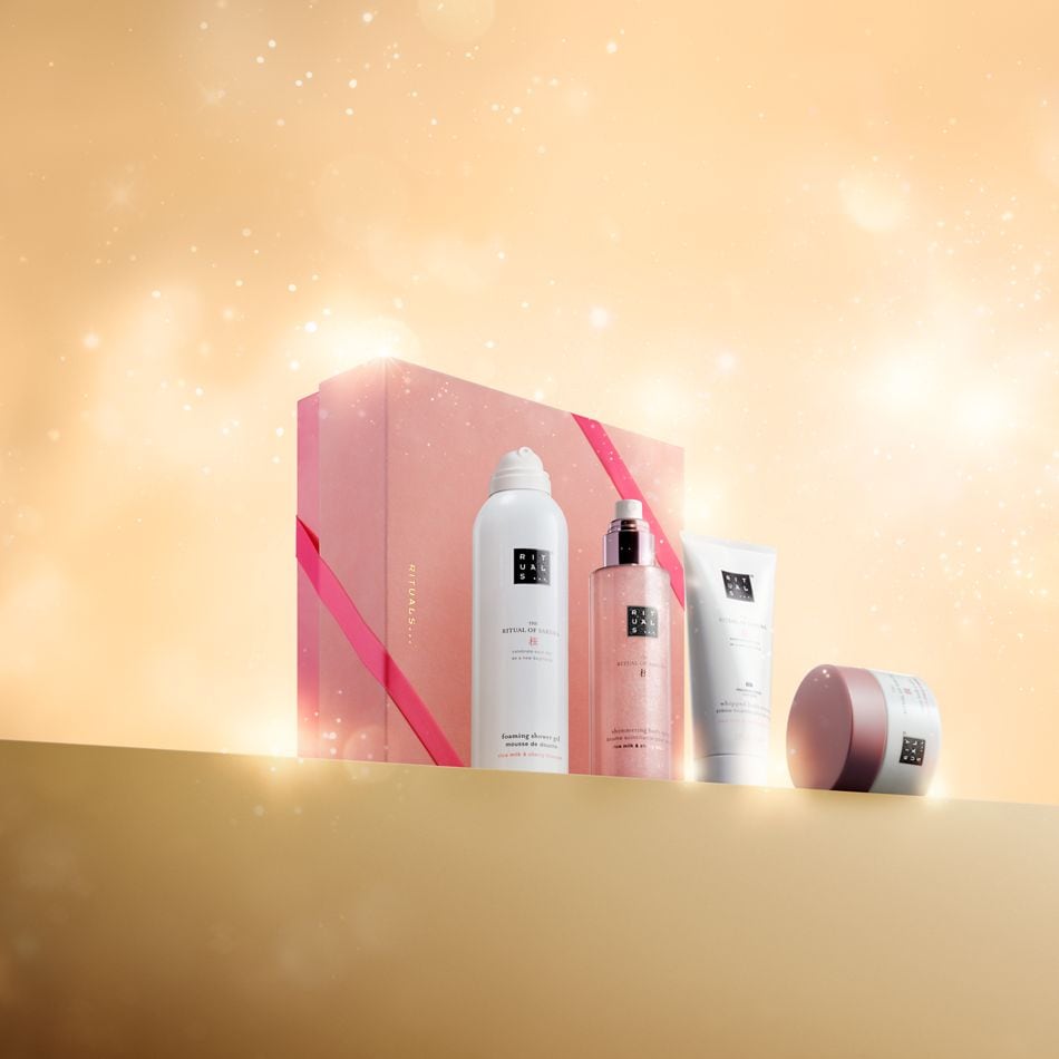 Das Rituals Geschenkset, bestehend aus einem rosa-weißem Duschschaum, einem schimmernden Körperöl, einer weißen Körpercreme und einem rosafarbenen Peeling, ist auf einem goldenen Podest vor einem glitzernden Hintergrund platziert.