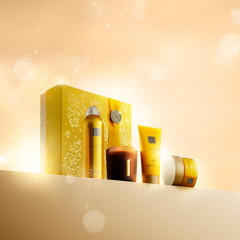Ein luxuriöses Geschenkset von Rituals in einer goldgelben Box mit weißem Blumenmuster, das ein Duschgel, eine Duftkerze, eine Körpercreme und eine Handcreme enthält, alles in passenden goldgelben Verpackungen.