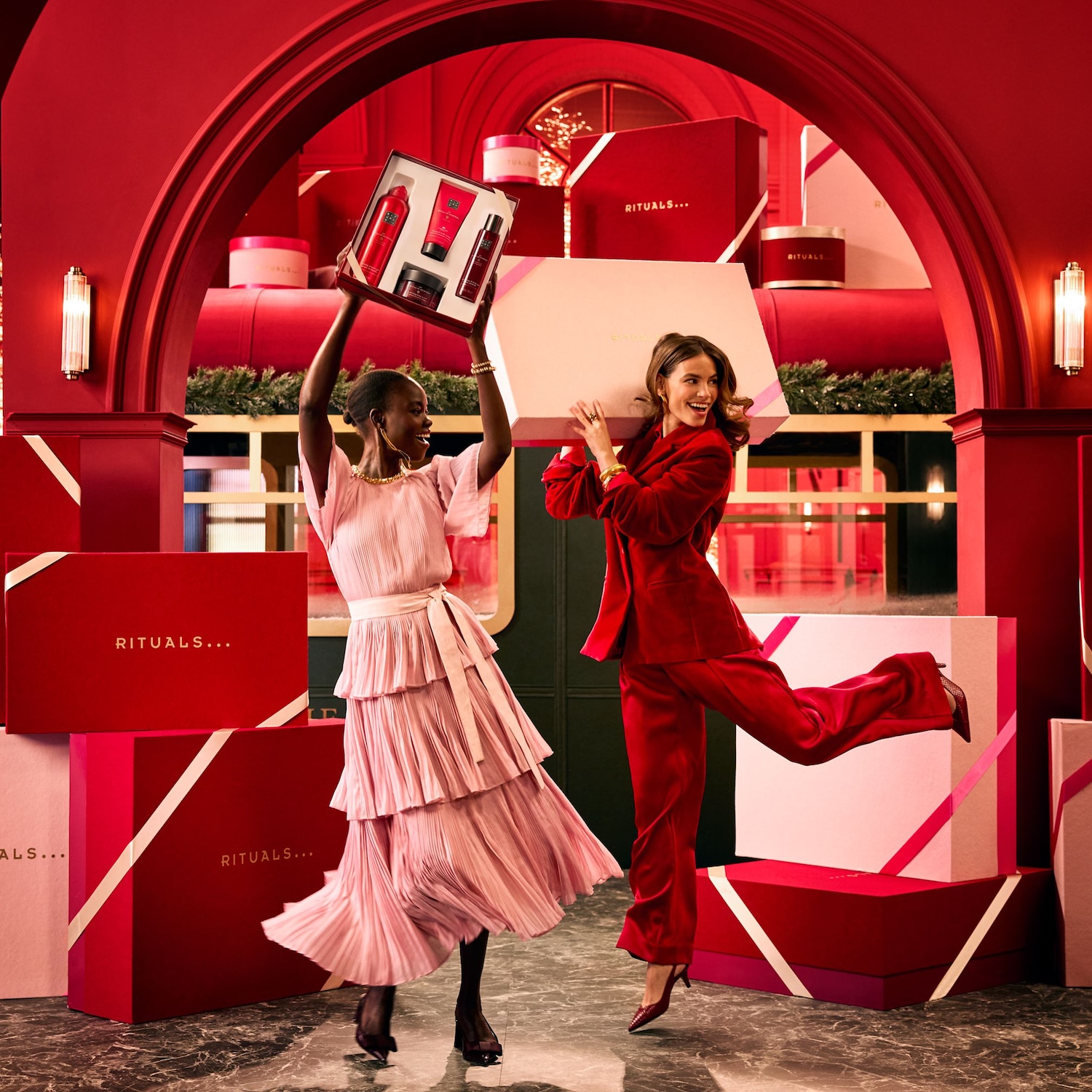 Zwei Frauen in festlicher Kleidung präsentieren Rituals-Geschenkboxen in einem eleganten Raum, der mit roten und weißen Geschenkpaketen dekoriert ist.