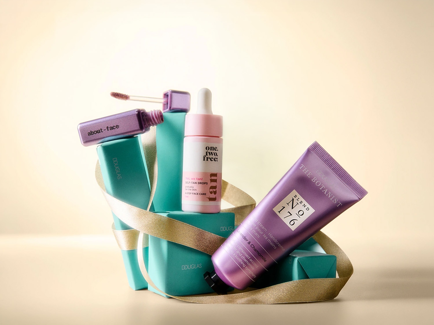 Een verzameling Douglas-producten, waaronder een paarse About-Face lipgloss, One.Two.Free! zelfbruinende druppels en The Botanist handcrème, allemaal gepresenteerd op turquoise dozen met een gouden lint.