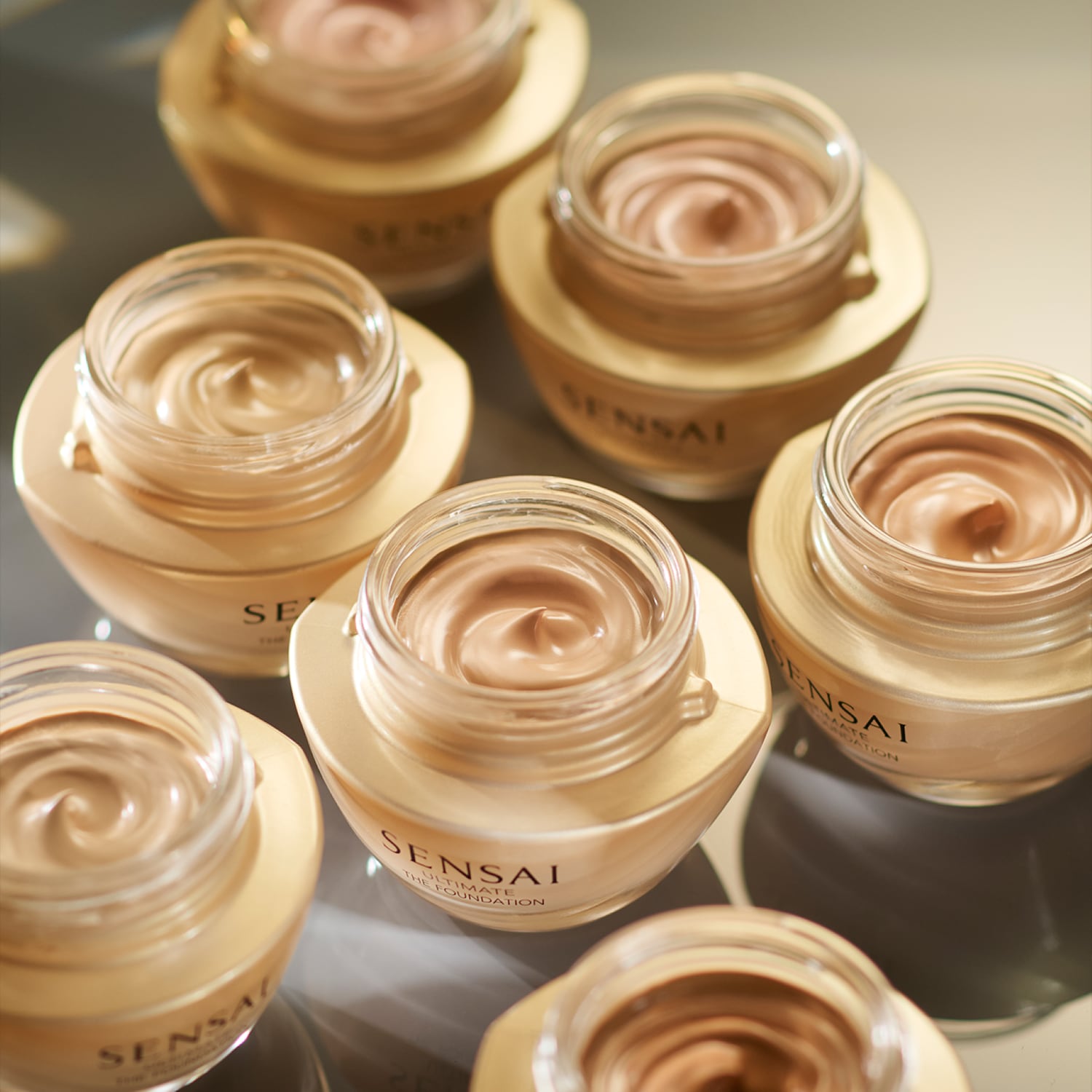 Découvrez la collection de fonds de teint Sensai Ultimate The Foundation, présentée dans des pots en verre transparent avec des bases dorées, offrant une gamme de nuances pour un teint parfait.