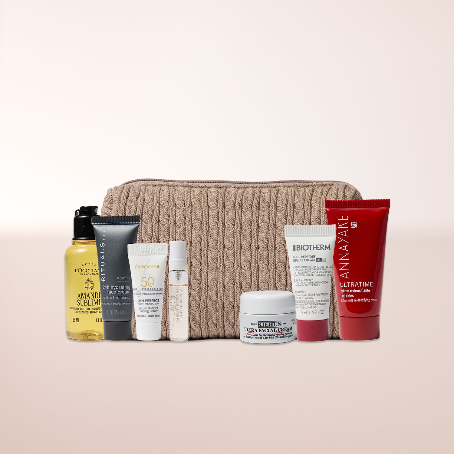 Ein beigefarbener, gestrickter Kulturbeutel ist mit einer Auswahl an Hautpflegeprodukten gefüllt, darunter eine gelbe Flasche L'Occitane Amande Sublime Duschöl, eine schwarze Tube Rituals Homme 24h Feuchtigkeitscreme, eine weiße Tube Lancaster Sun Perfect Youth Protection Serum, ein kleines Fläschchen Issey Miyake Lumière D'Issey Parfüm, ein weißer Tiegel Kiehl's Ultra Facial Cream, eine weiße Tube Biotherm Blue Peptides Uplift Cream SPF 30 und eine rote Tube Annayake Ultratime Anti-Falten-Creme.