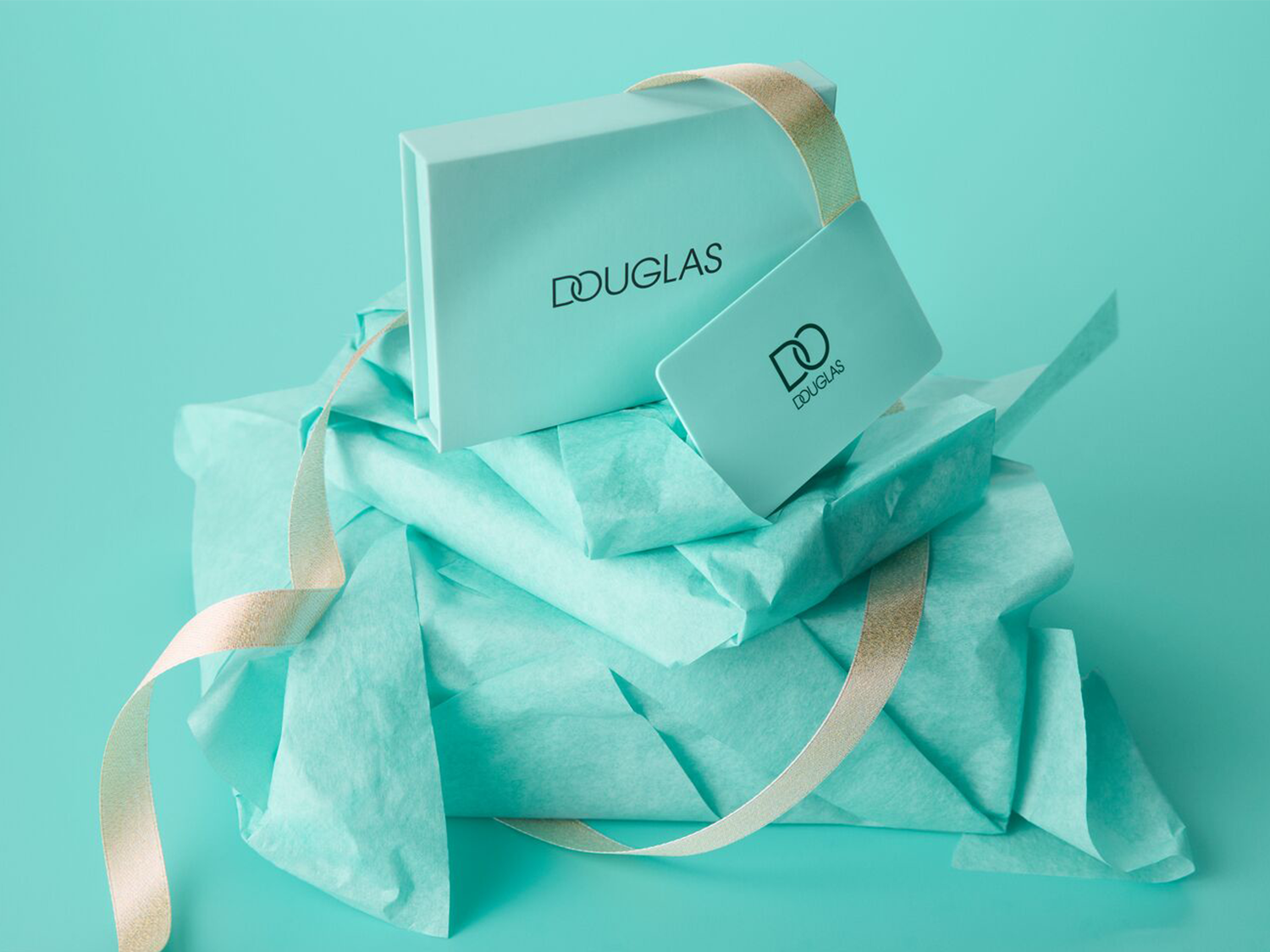 Un coffret cadeau Douglas bleu sarcelle avec un ruban doré, posé sur du papier de soie froissé de la même couleur, avec une carte cadeau assortie sur le dessus.