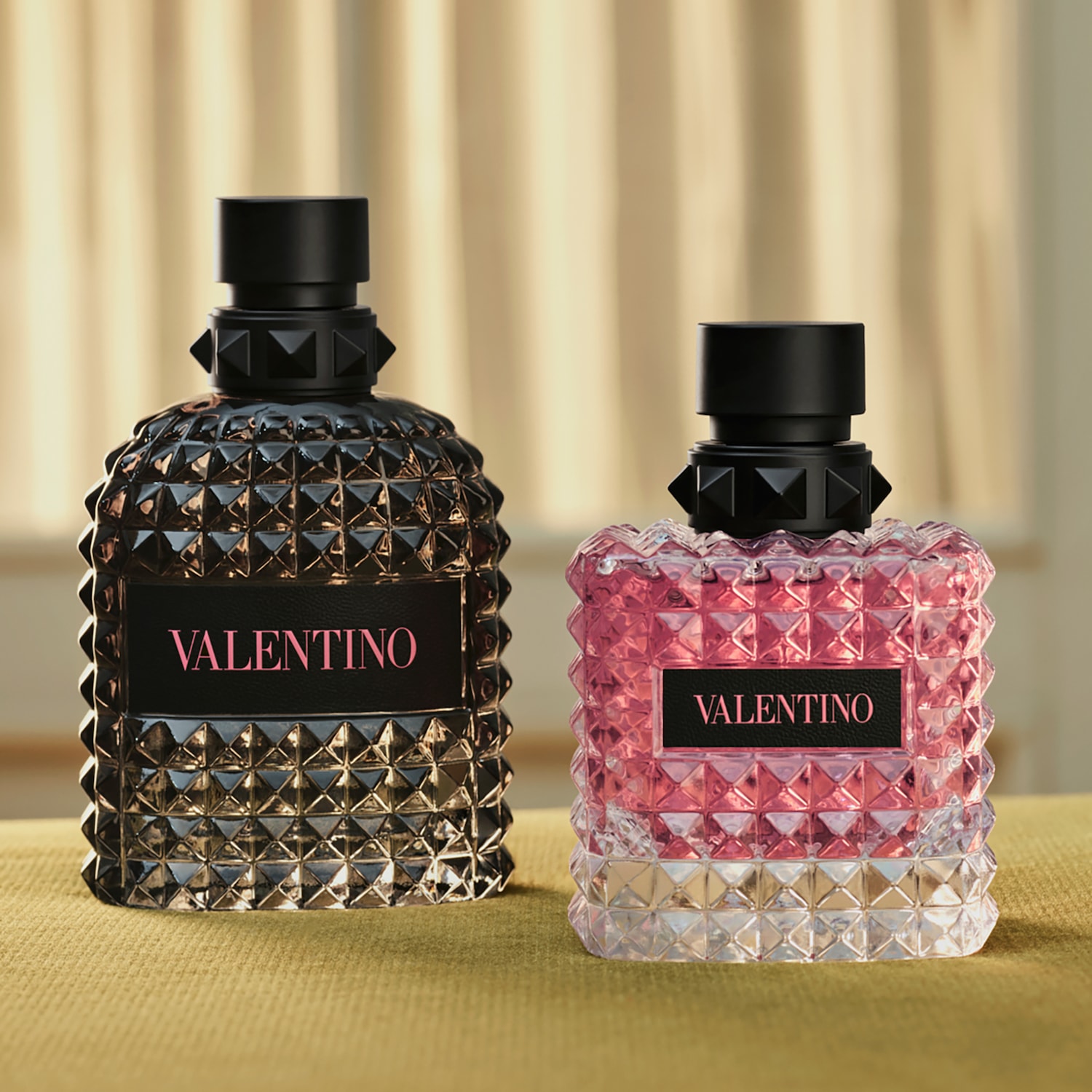 Zwei Valentino Born in Roma Parfümflaschen stehen nebeneinander, eine in dunklem Glas mit schwarzem Inhalt und die andere in klarem Glas mit rosafarbenem Inhalt, beide mit einem schwarzen Etikett mit dem Namen „VALENTINO“ in Rosa.