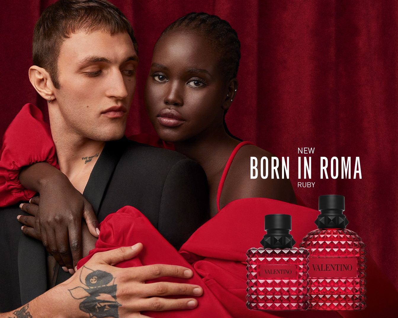 Ein Mann und eine Frau posieren vor einem roten Hintergrund, die Frau umarmt den Mann, während zwei Flaschen des neuen Parfüms "Born in Roma Ruby" von Valentino im Vordergrund stehen.