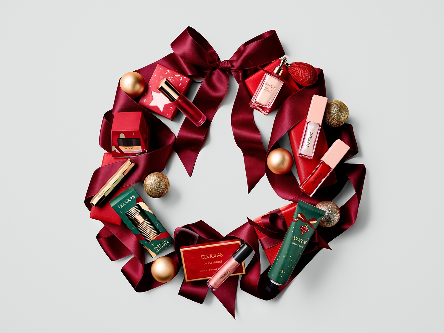 Découvrez une couronne de Noël festive ornée d'un ruban rouge satiné, de boules dorées scintillantes et d'une sélection de produits de beauté Douglas, incluant des rouges à lèvres, des parfums et des crèmes pour les mains, parfaits pour les fêtes.