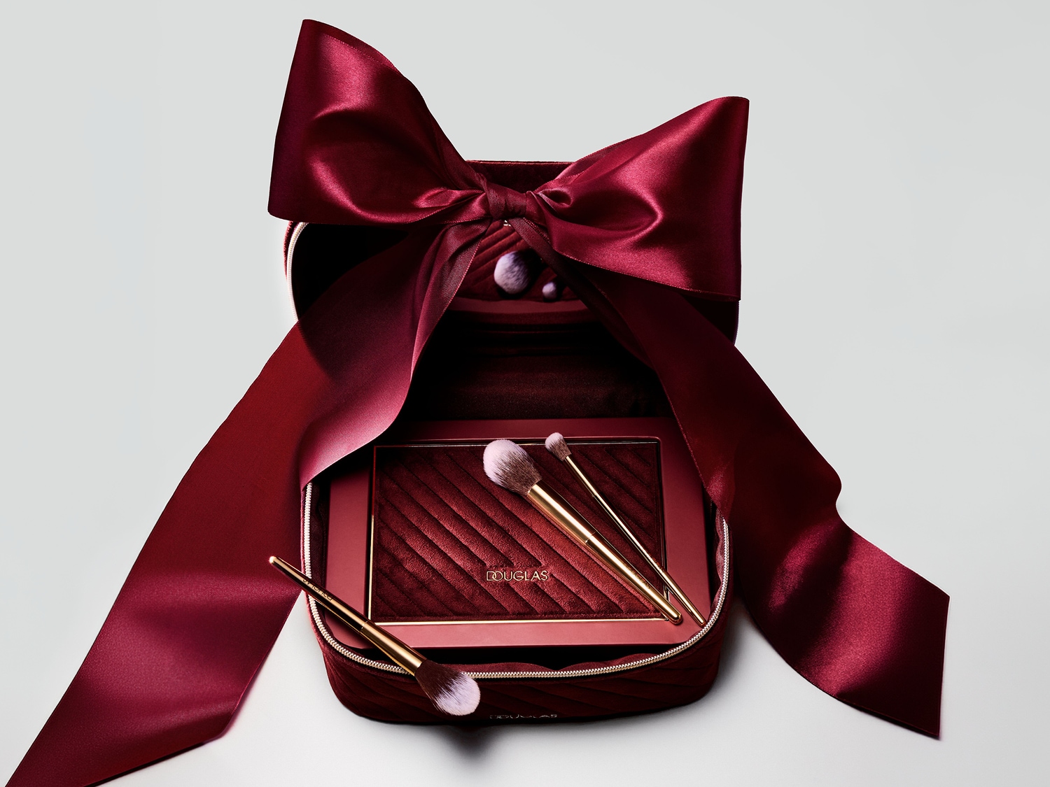 Un coffret de maquillage Douglas rouge bordeaux ouvert, orné d'un grand ruban de satin de la même couleur, révèle une palette de fards à paupières et trois pinceaux de maquillage dorés.