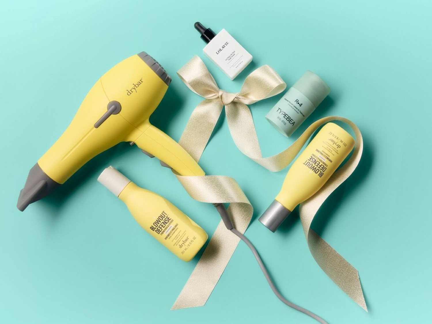 Un asciugacapelli giallo Drybar, due flaconi gialli di Blowout Defense, un flacone verde di Typebea R.4 e un flacone bianco di Lolavie, tutti disposti su uno sfondo blu acqua con un nastro dorato.