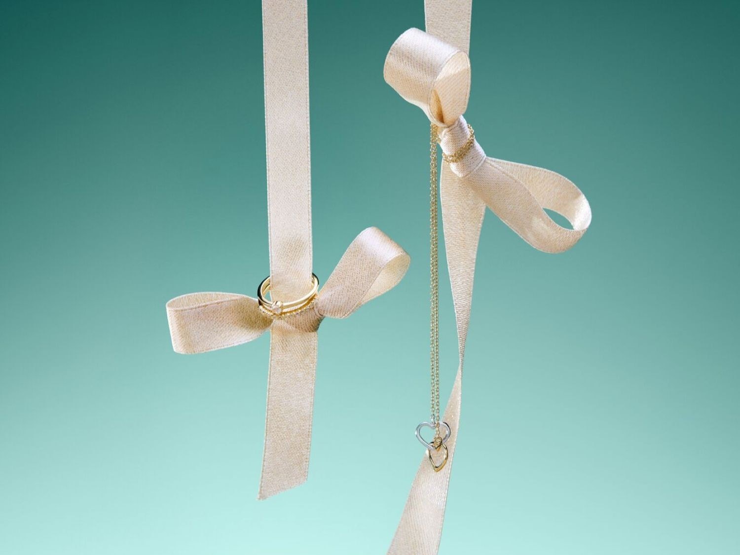 Un anello d'oro e una collana con ciondolo a forma di cuore pendono da nastri color crema su uno sfondo verde acqua, simboleggiando eleganza e amore.