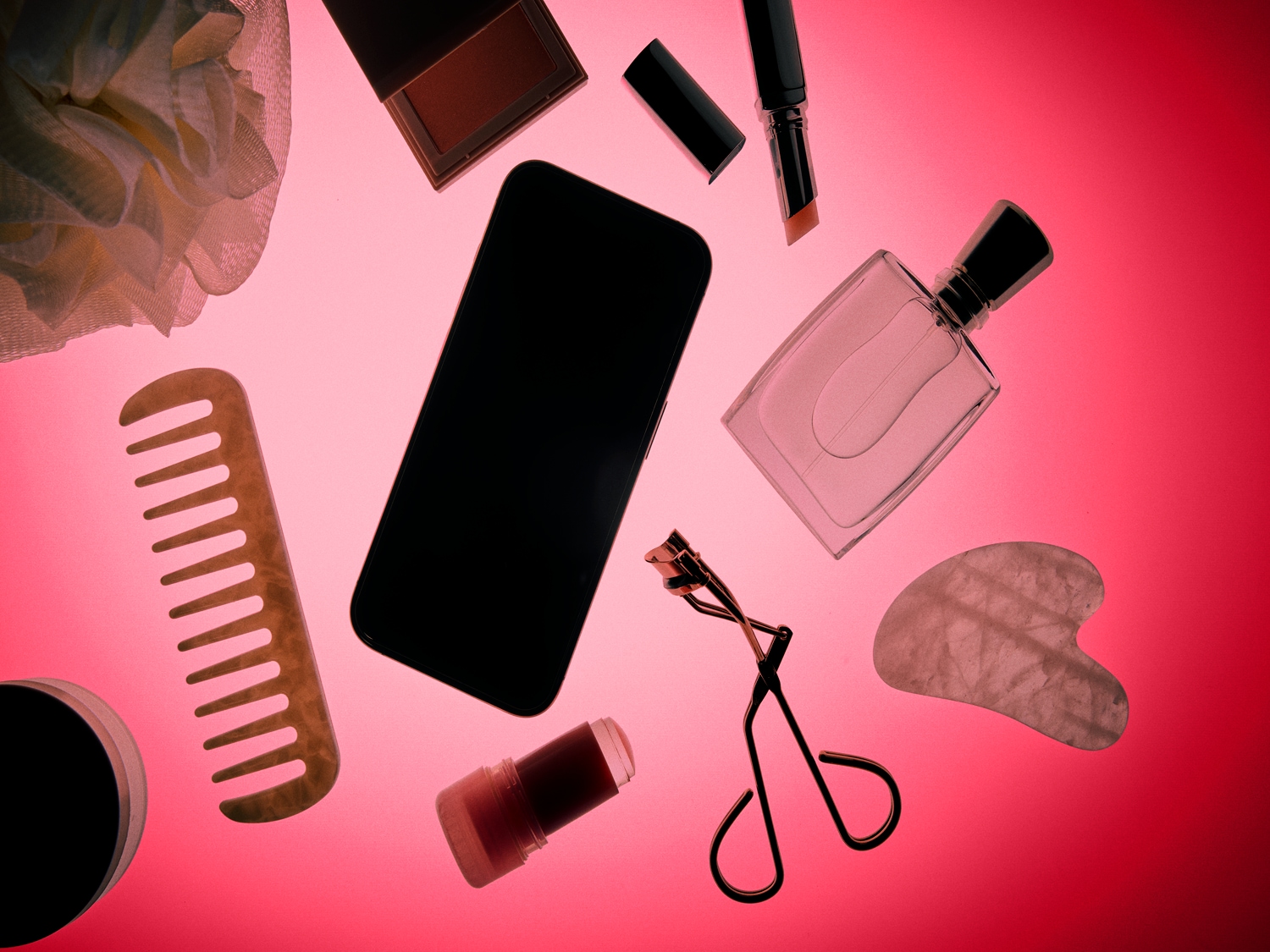 Een verzameling van beautyproducten en accessoires, waaronder een mobiele telefoon, parfumflesje, make-up, kam, wimperkruller, gua sha steen en een badspons, allemaal verspreid over een roze achtergrond.