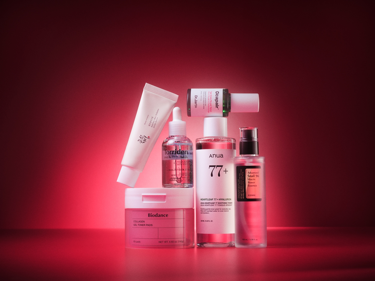 Een verzameling van zes Koreaanse huidverzorgingsproducten, waaronder een zonnebrandcrème, serum, toner, essence en collageen gel toner pads, staat op een levendige rode achtergrond.
