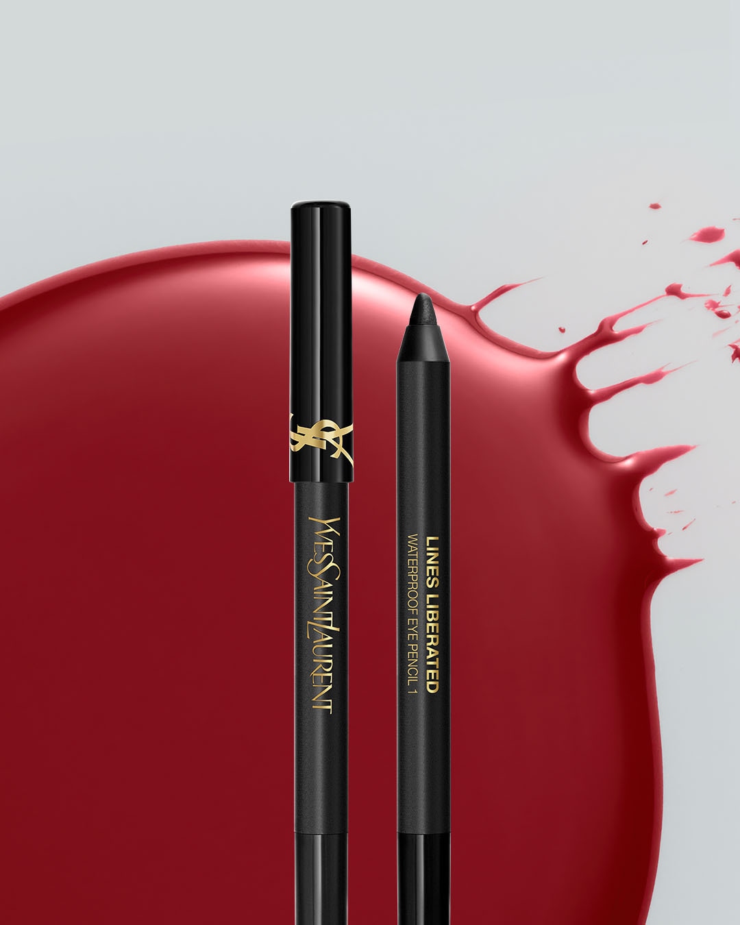 Dwa czarne wodoodporne kredki do oczu Yves Saint Laurent, jedna z logo marki, a druga z napisem "LINES LIBERATED WATERPROOF EYE PENCIL 1", leżą na tle czerwonej, błyszczącej plamy.