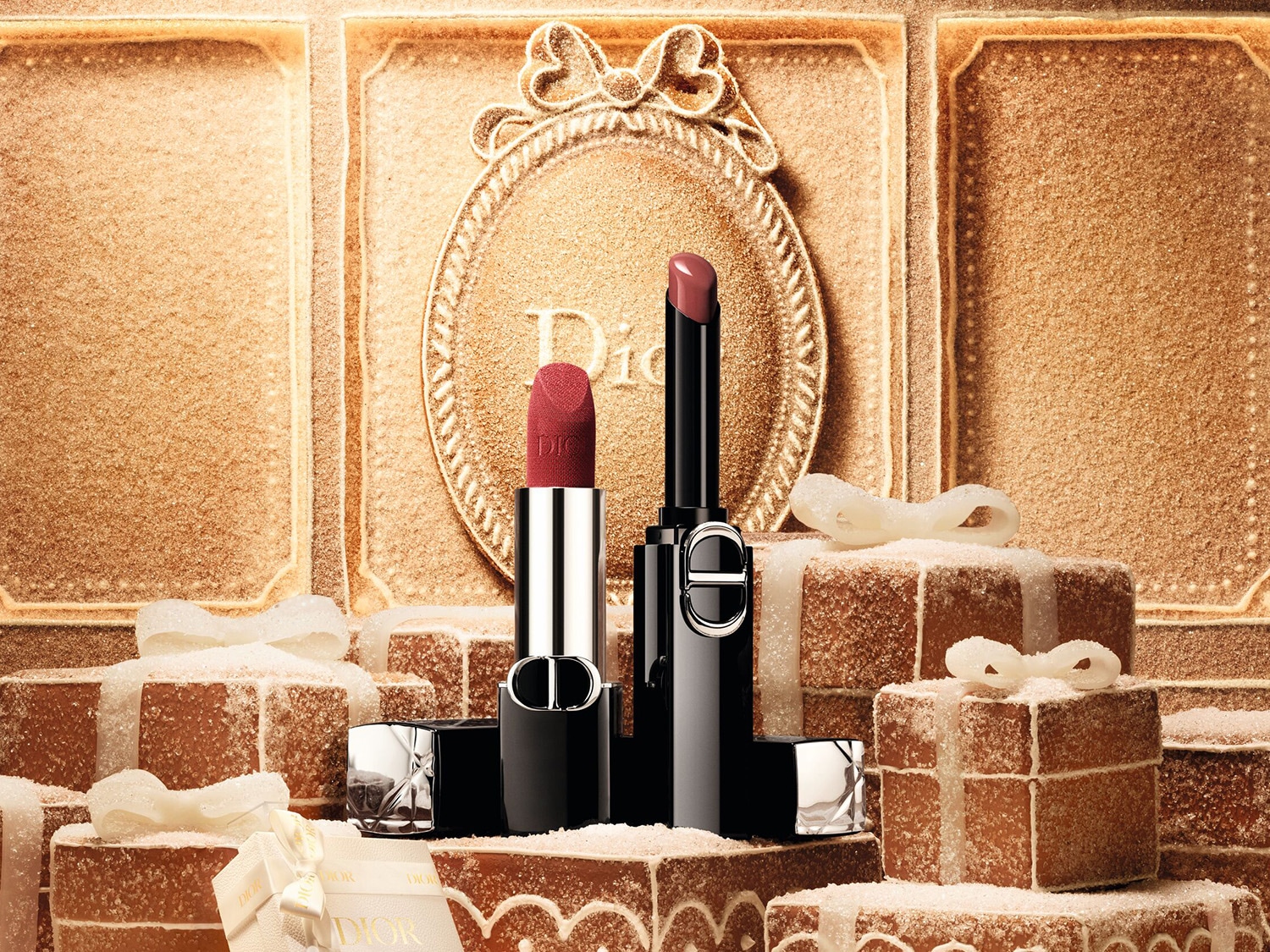 Deux rouges à lèvres Dior, l'un rouge mat et l'autre rose brillant, sont présentés au milieu de cadeaux de Noël dorés et scintillants, créant une ambiance festive et élégante.