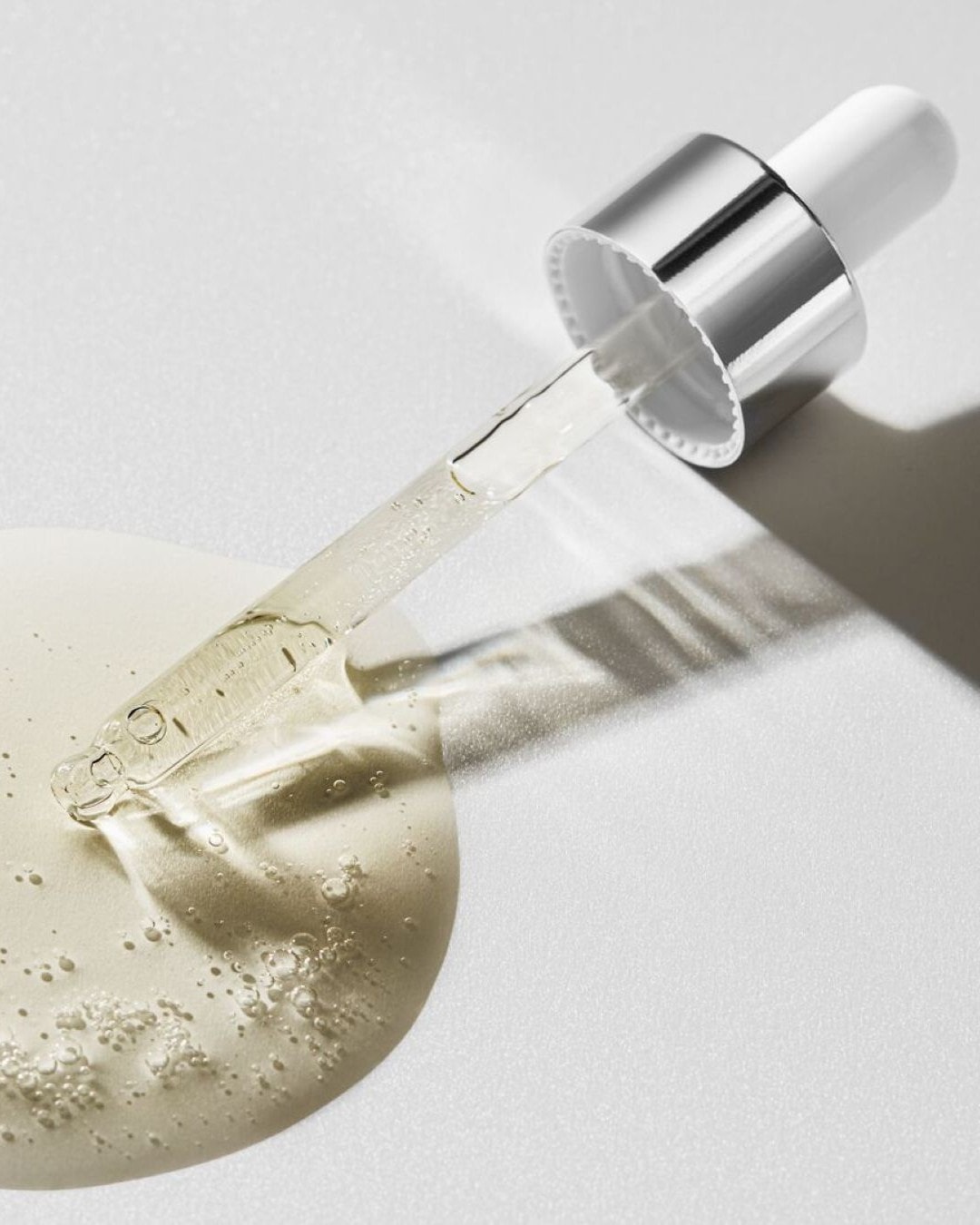 Un contagocce d'argento e bianco rilascia un siero trasparente e dorato su una superficie bianca, con bolle visibili nel liquido.