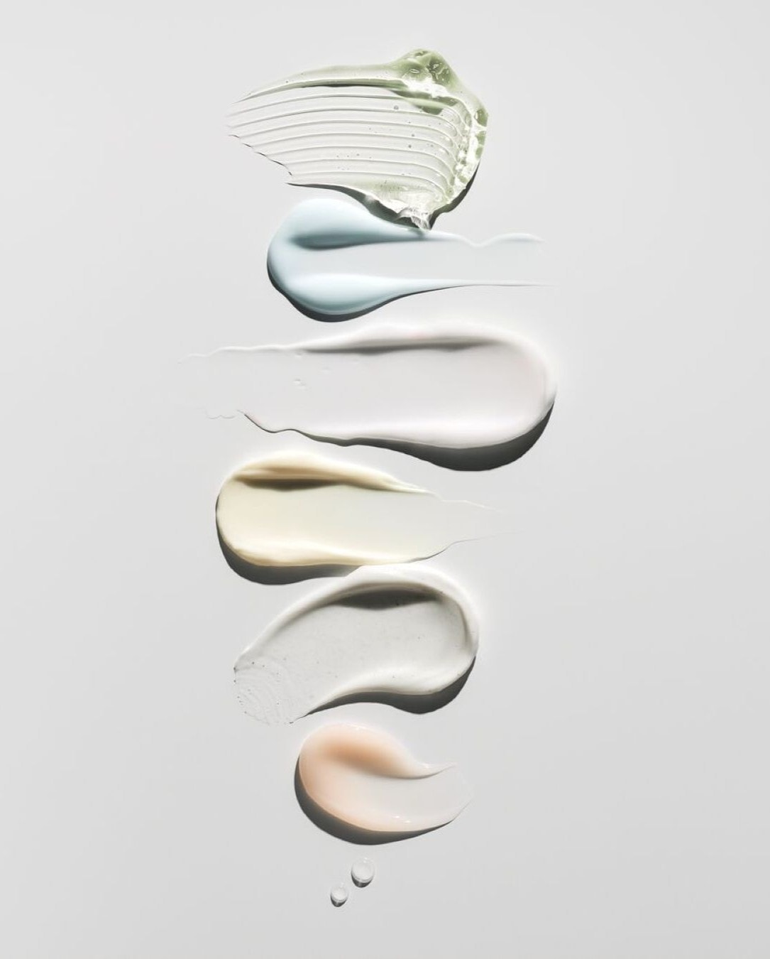 Una serie di campioni di prodotti di bellezza, tra cui un gel verde chiaro, una crema azzurra, una crema bianca, una crema beige, una crema grigia e una crema color pesca, sono disposti verticalmente su uno sfondo bianco.