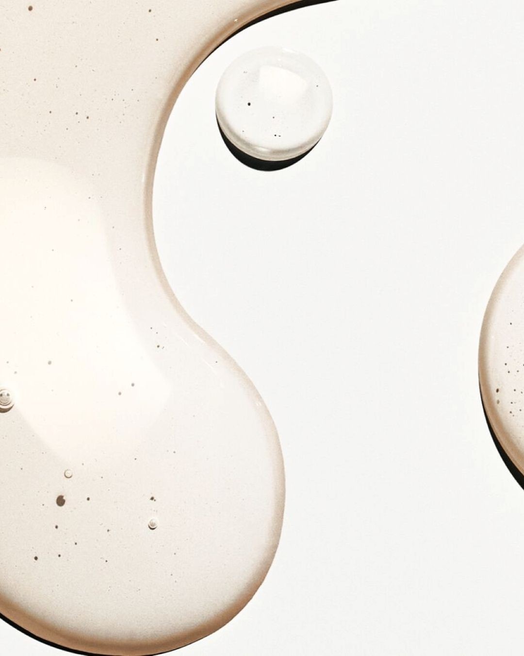 Una texture chiara e liquida con piccole bolle e puntini neri si estende su uno sfondo bianco, con una goccia più piccola e rotonda in alto a destra e una forma ovale più grande in basso a sinistra.