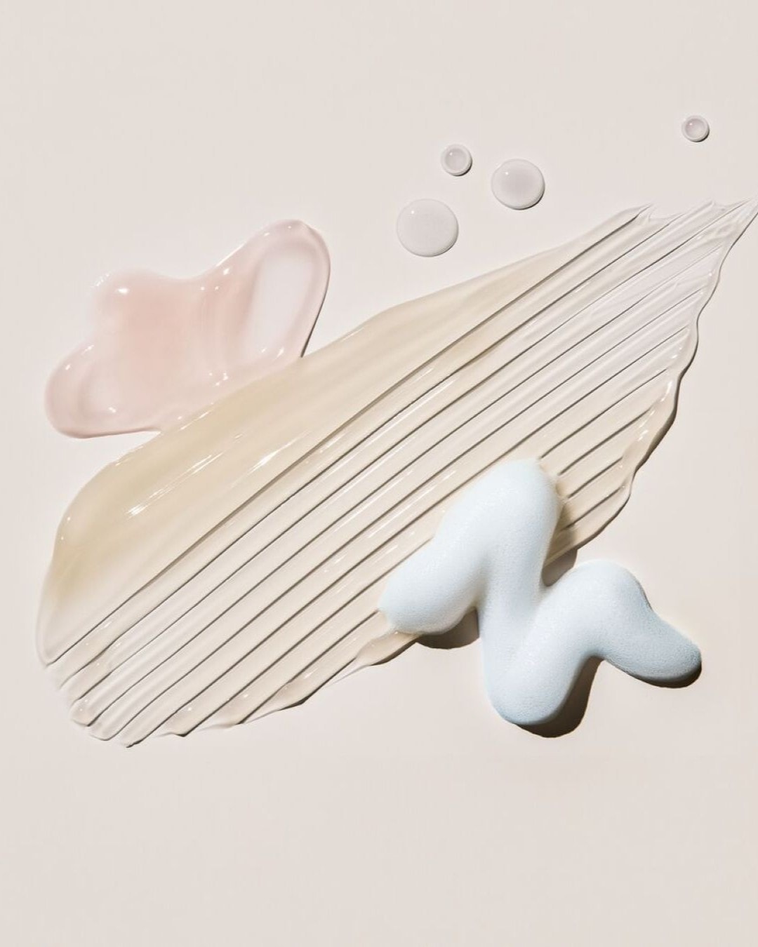 Una varietà di texture cosmetiche, tra cui un gel rosa chiaro, una crema beige spalmata, goccioline trasparenti e una schiuma bianca a forma di S, sono disposte su uno sfondo neutro.