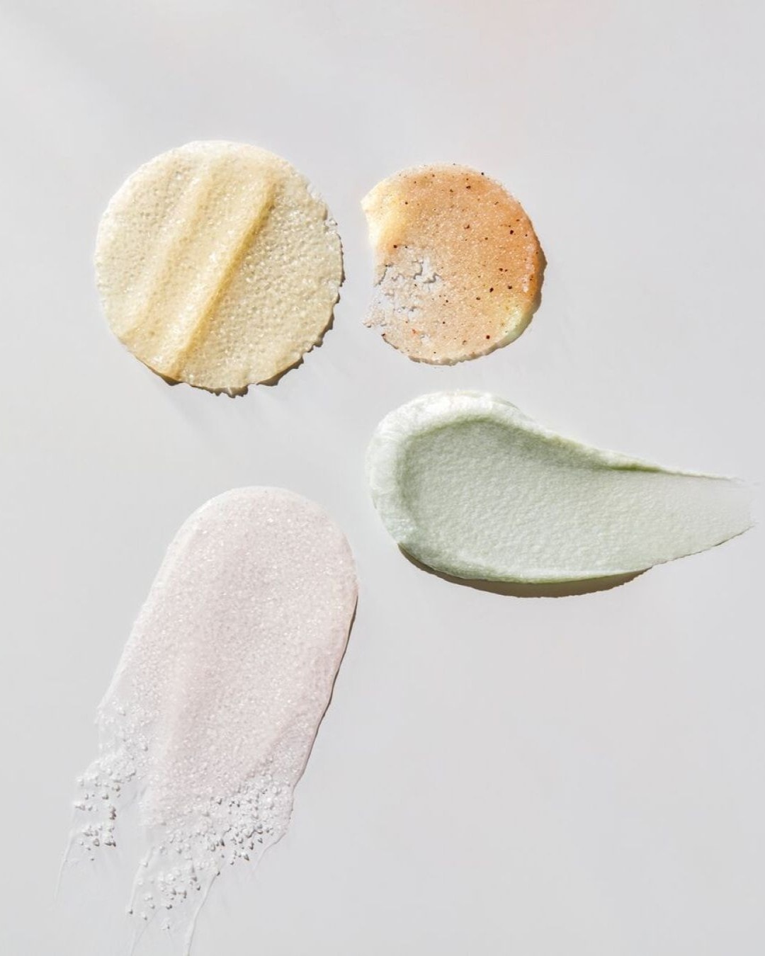 Quattro campioni di scrub per il corpo sono disposti su uno sfondo bianco, due a forma circolare e due a striscia, con texture e colori diversi che vanno dal beige chiaro al verde menta e al rosa pallido.
